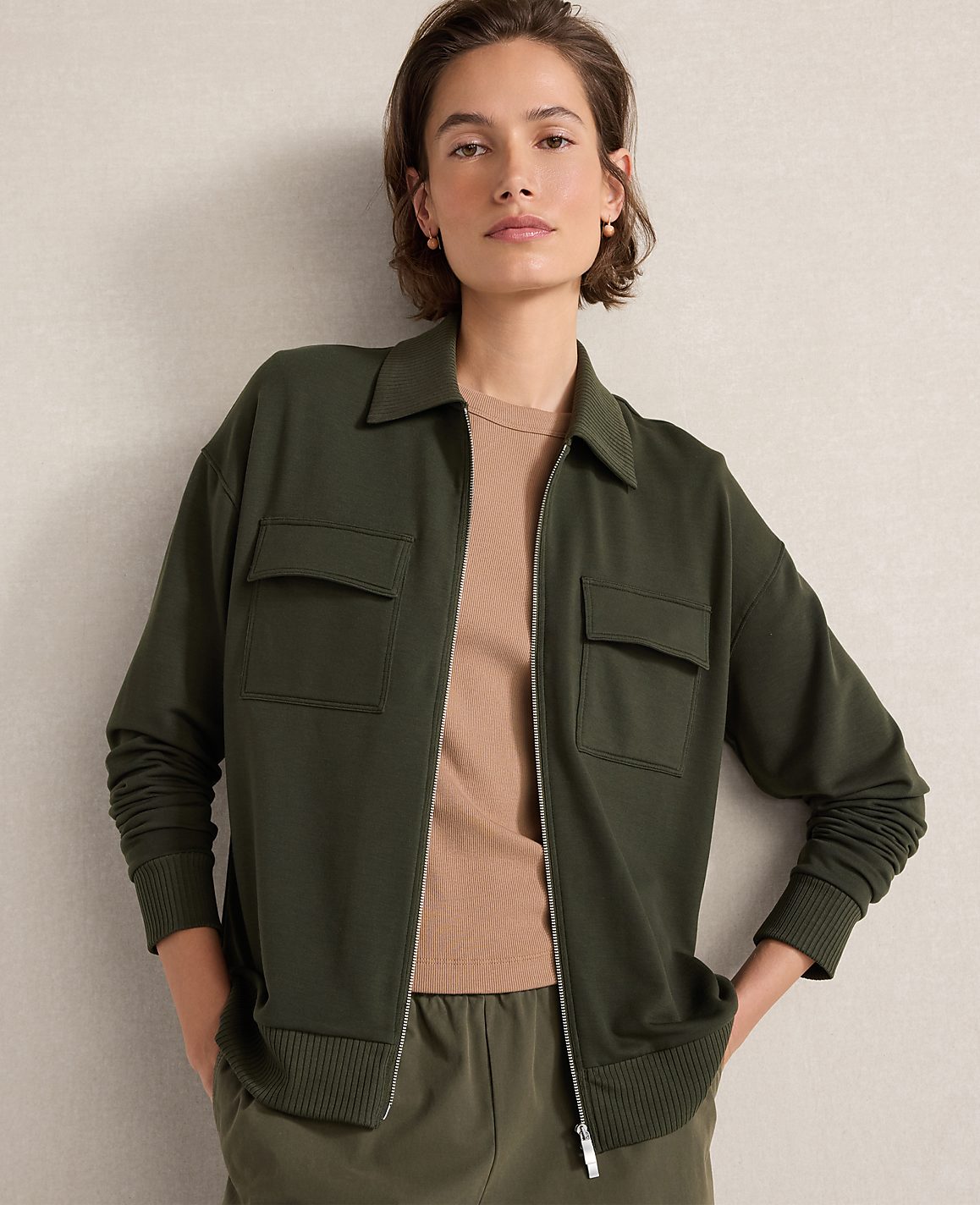 A woman in a green zip-up jacket and tan shirt stands against a plain, light-colored background.