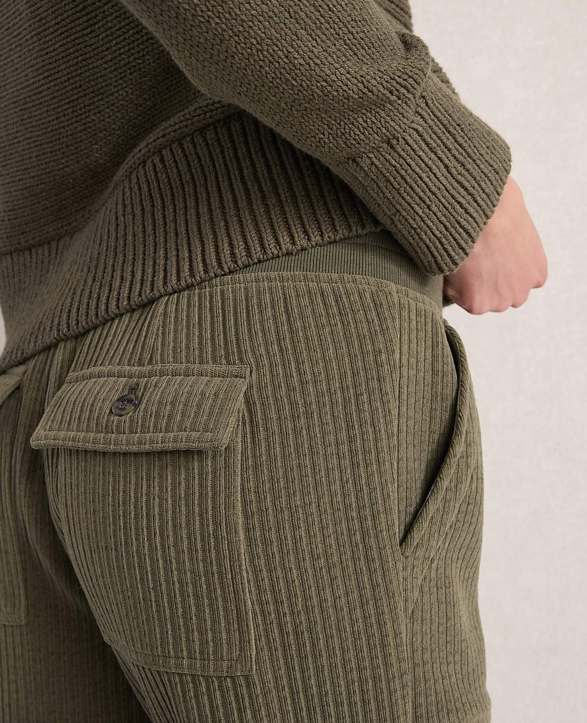 A person's back is shown wearing a green ribbed sweater and corduroy pants with a buttoned pocket.