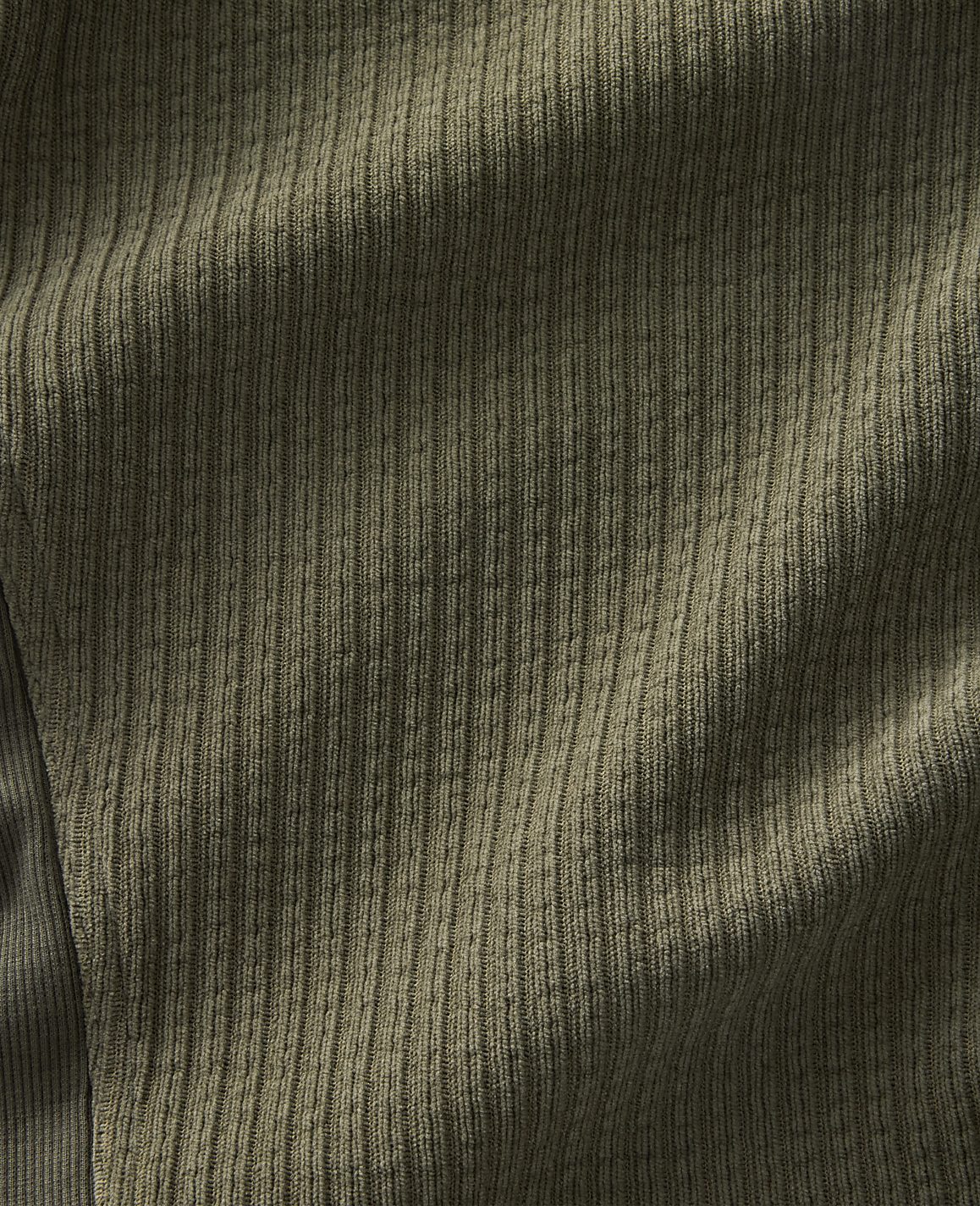 A close-up view of textured olive-green fabric with soft shadows creating depth in the foreground, against a subtly darker, ribbed background.