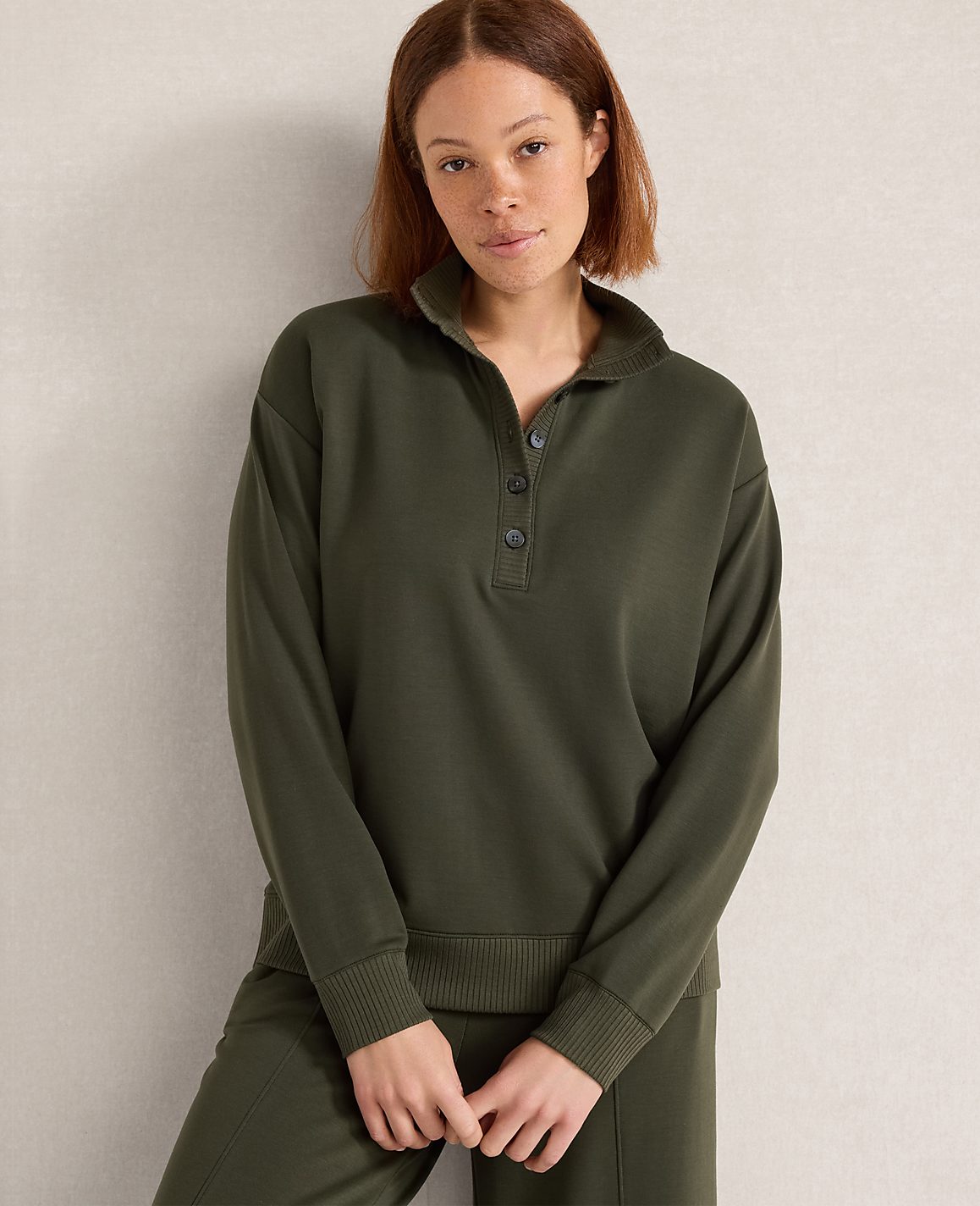 A woman in a green collared sweatshirt and matching pants stands against a plain, light-colored wall.