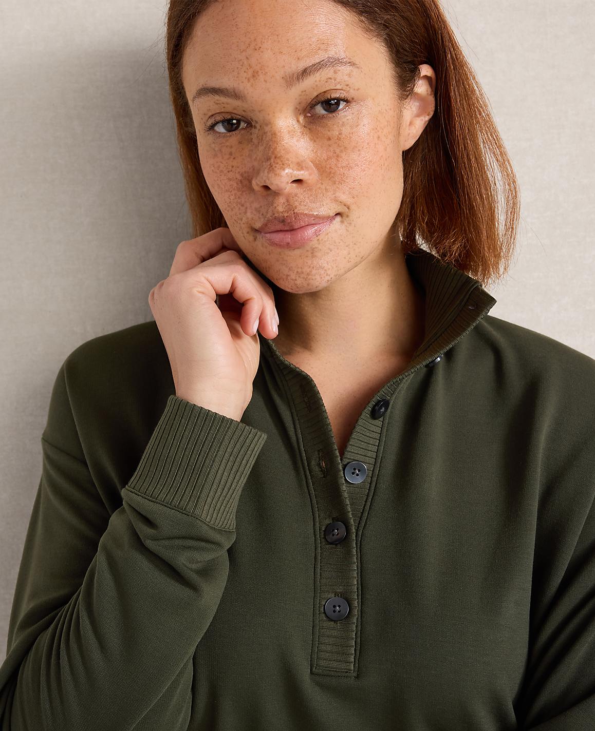 A woman with freckles and short brown hair, wearing a green collared sweatshirt, is looking towards the camera with her hand near her face, against a plain, light-colored background.