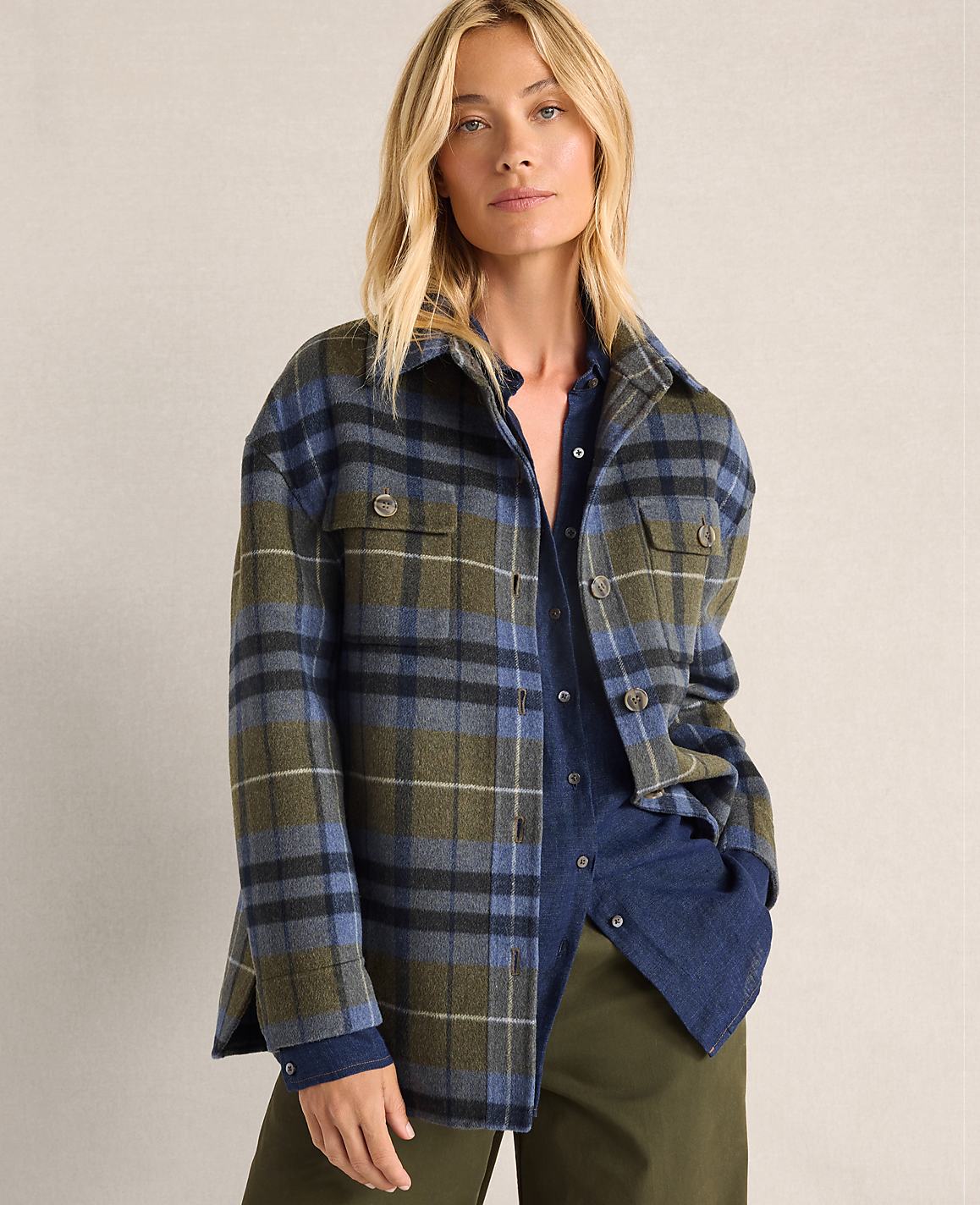 A blonde woman in a plaid jacket and dark shirt stands against a plain, light-colored background.