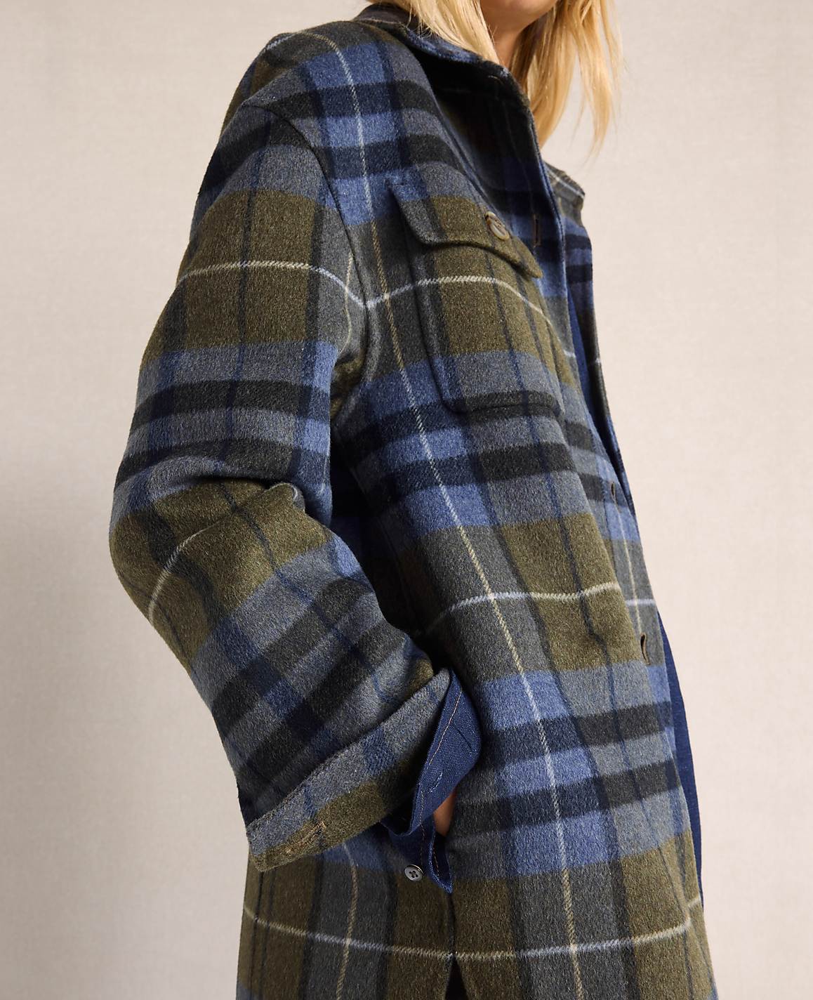 A person in a blue and green plaid jacket stands against a plain, light-colored background.