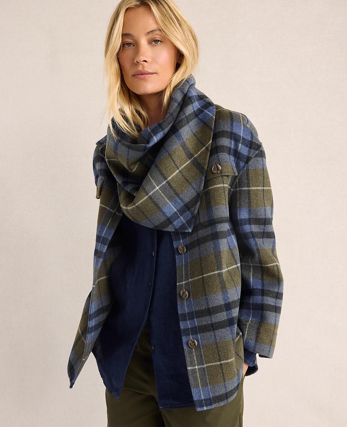 A blonde woman in a plaid jacket and scarf stands against a plain background.