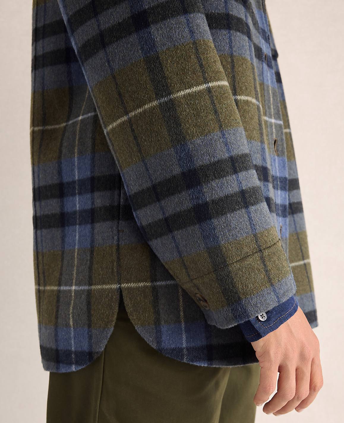 A person in a plaid shirt and olive pants stands against a plain background.