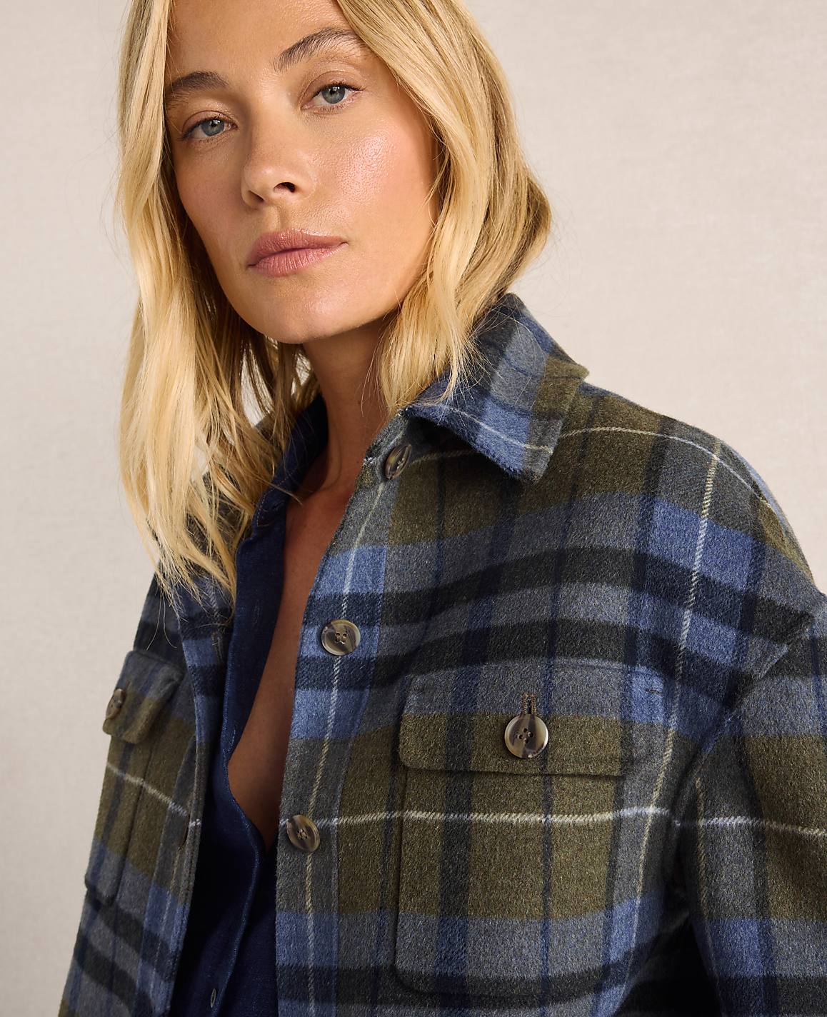 A woman with blonde hair and fair skin wears a blue and green plaid jacket in the foreground, with a neutral, textured background.