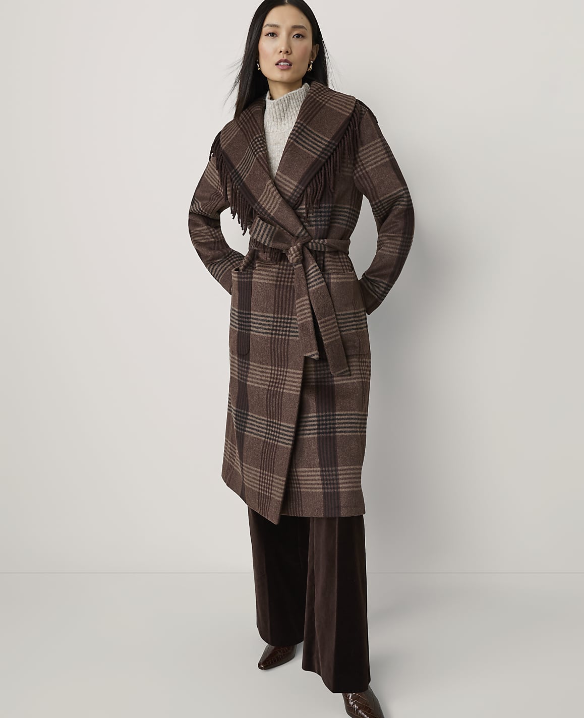 A woman in a brown plaid coat with fringe stands against a plain white background.
