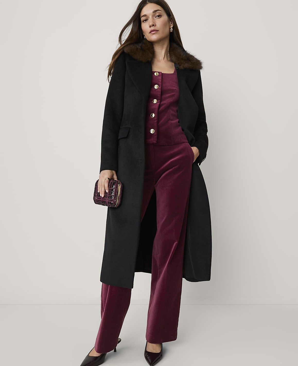 A woman in a black coat and burgundy velvet jumpsuit stands against a plain, light background, holding a small clutch.