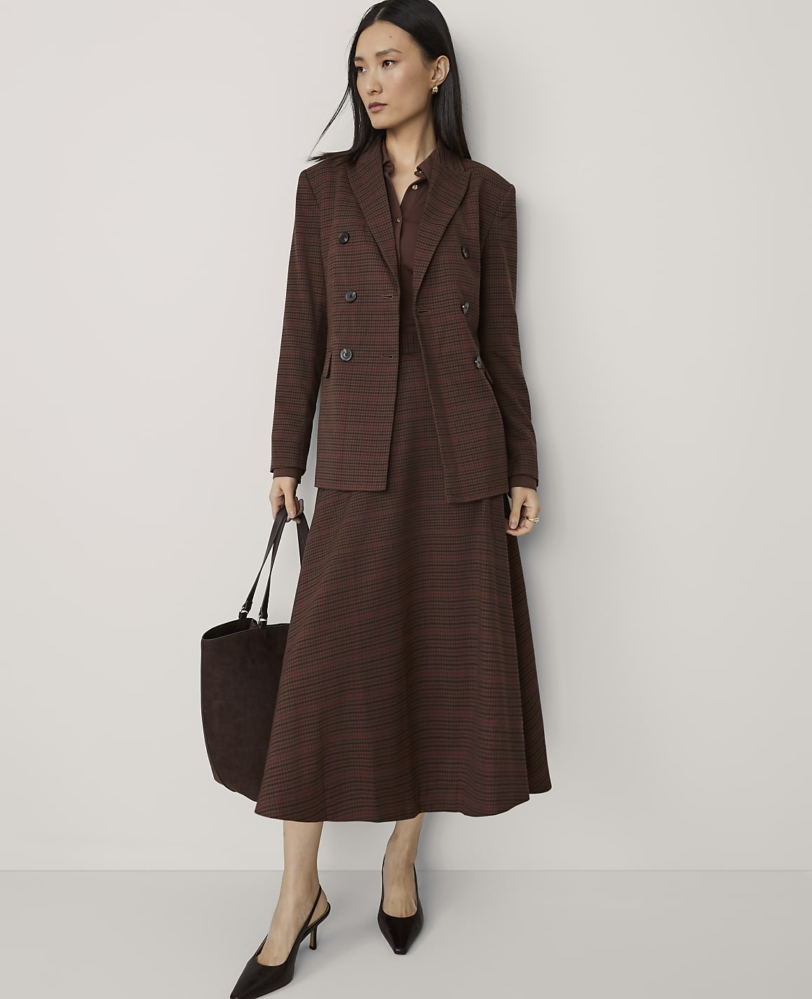 A woman in a brown plaid suit and heels stands with a tote bag against a plain, light-colored wall.
