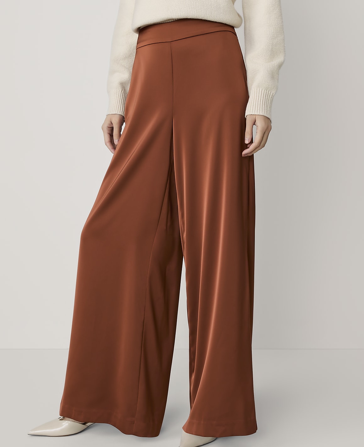 A person wearing rust-colored wide-leg pants and a cream sweater stands against a plain white background.