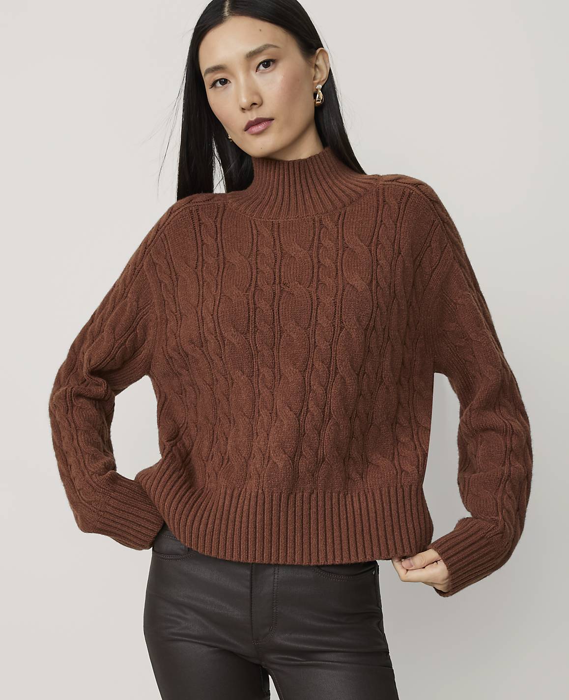 A woman in a brown cable-knit turtleneck sweater and dark pants poses against a plain white background.