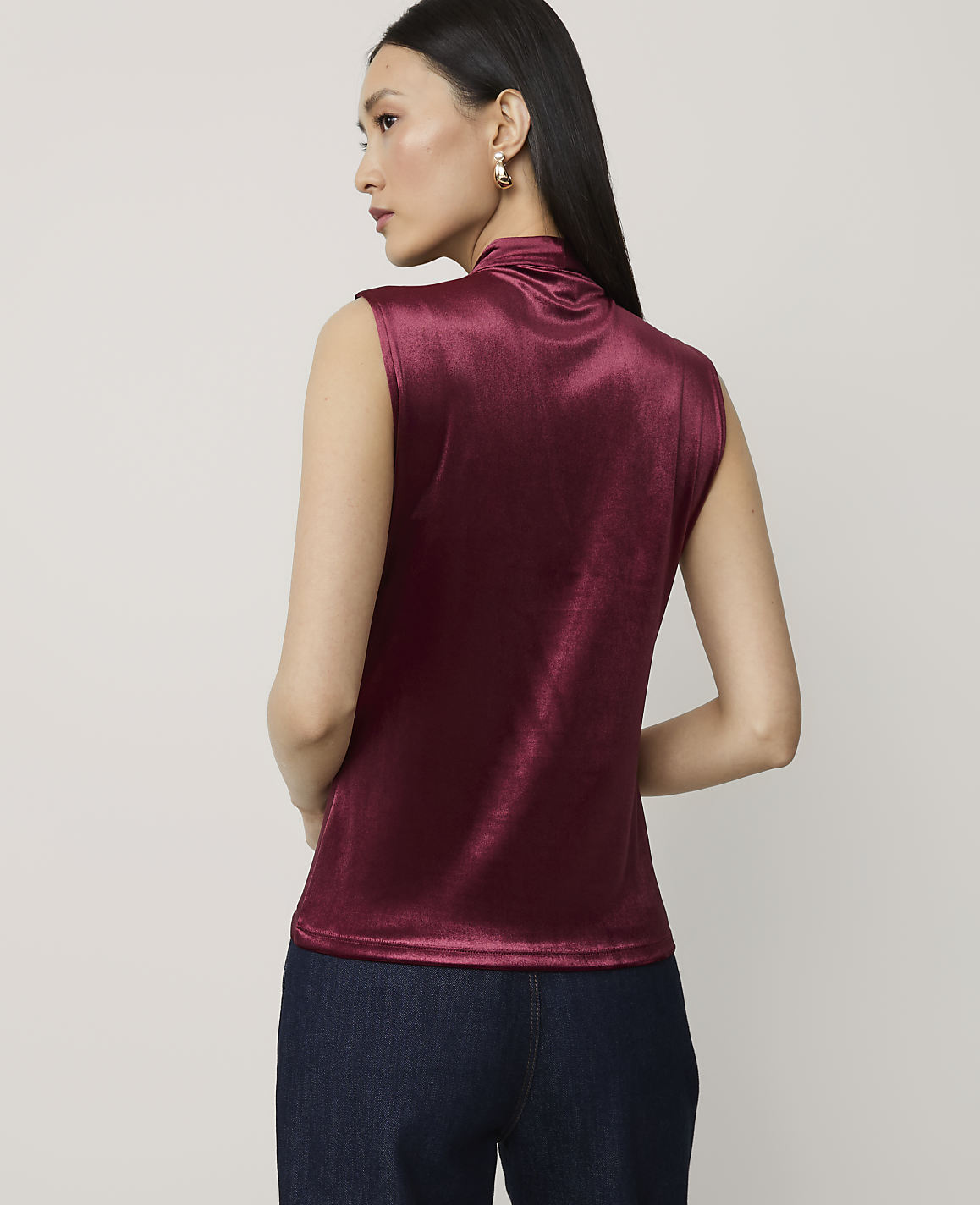 A woman in a burgundy velvet top and dark jeans stands against a plain, light gray background.