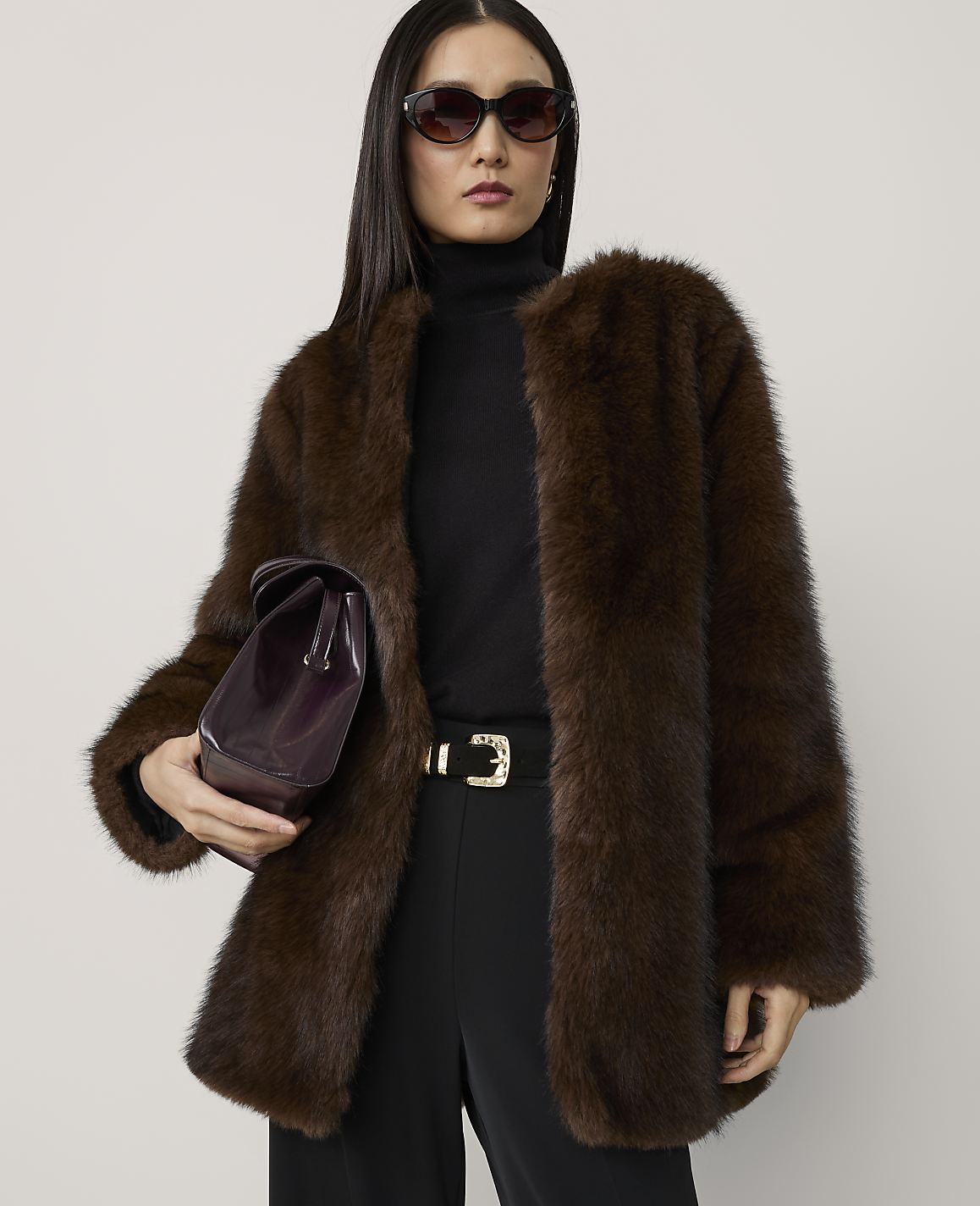 A woman in a luxurious brown fur coat and black turtleneck stands against a plain, light gray background, holding a dark purple clutch.