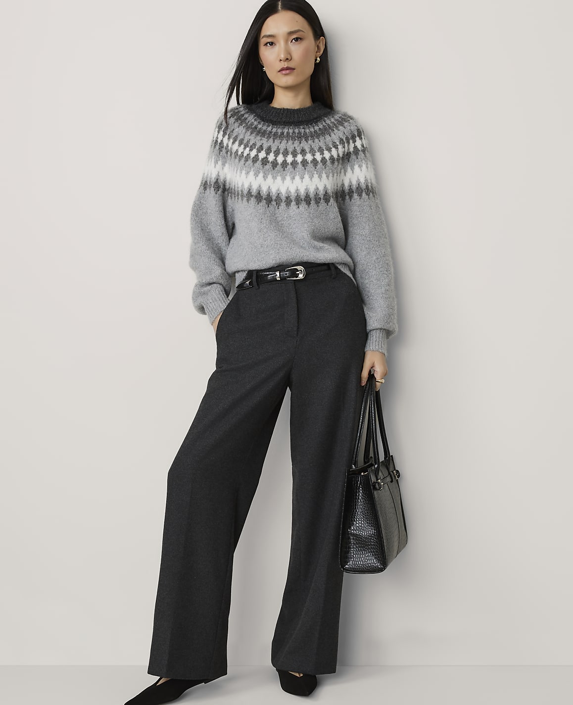 A woman in a grey fair isle sweater and wide-leg trousers stands against a plain white background, holding a black handbag.