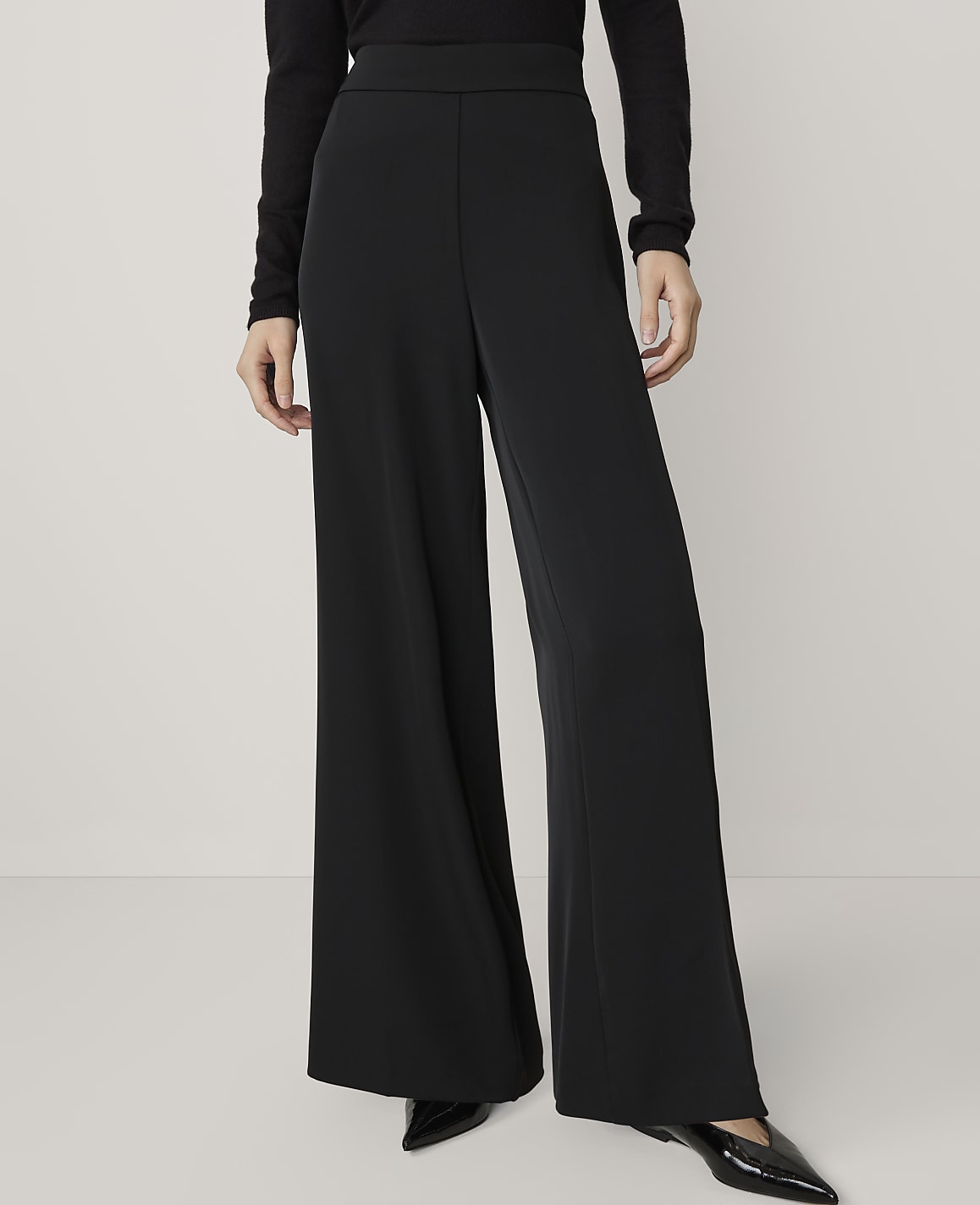 A person wearing black wide-leg pants and black pointed-toe shoes stands against a plain white background.