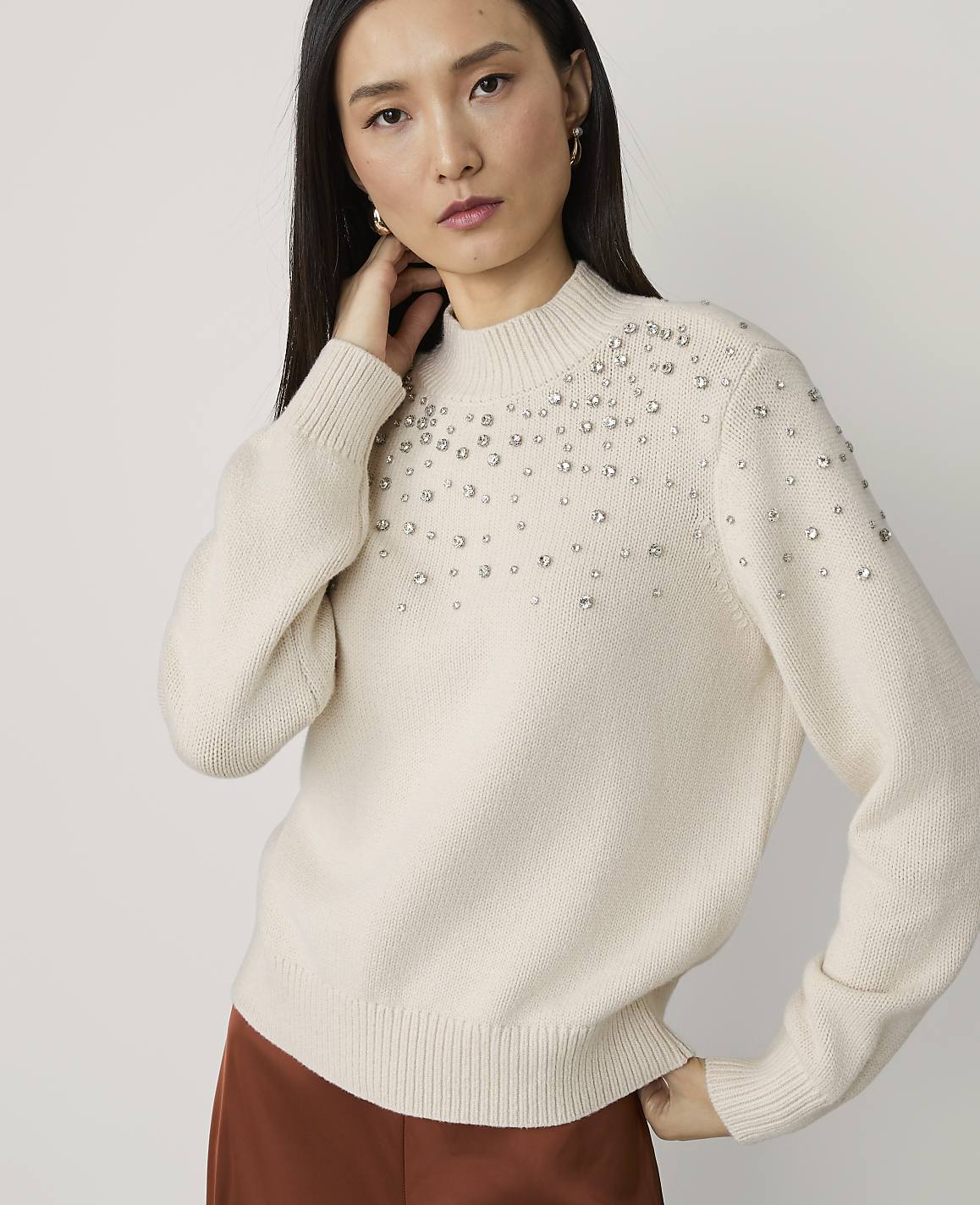 A woman in a cream-colored sweater adorned with rhinestones poses against a plain white background.