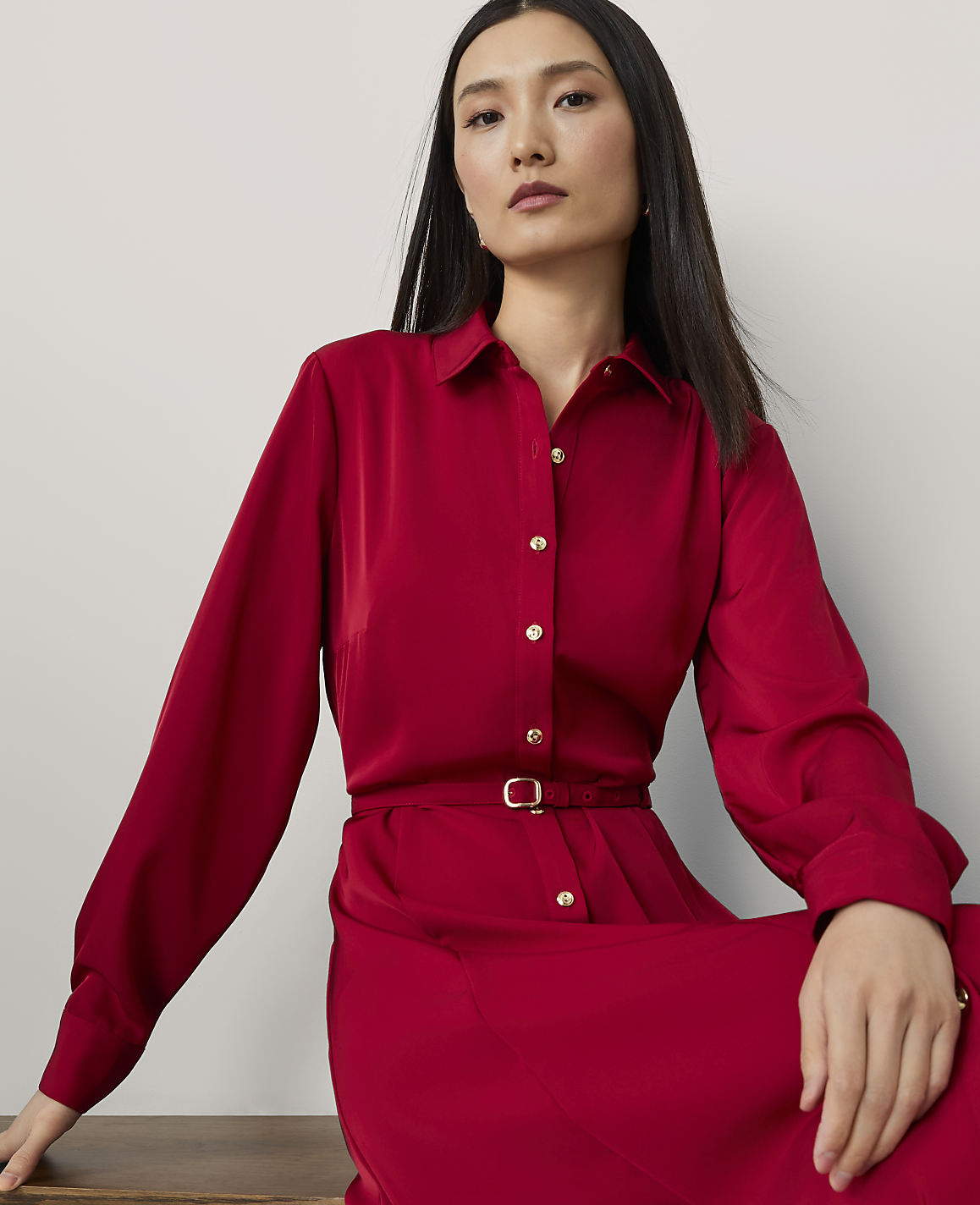 A woman in a red dress with a belted waist is seated against a plain, light-colored background.