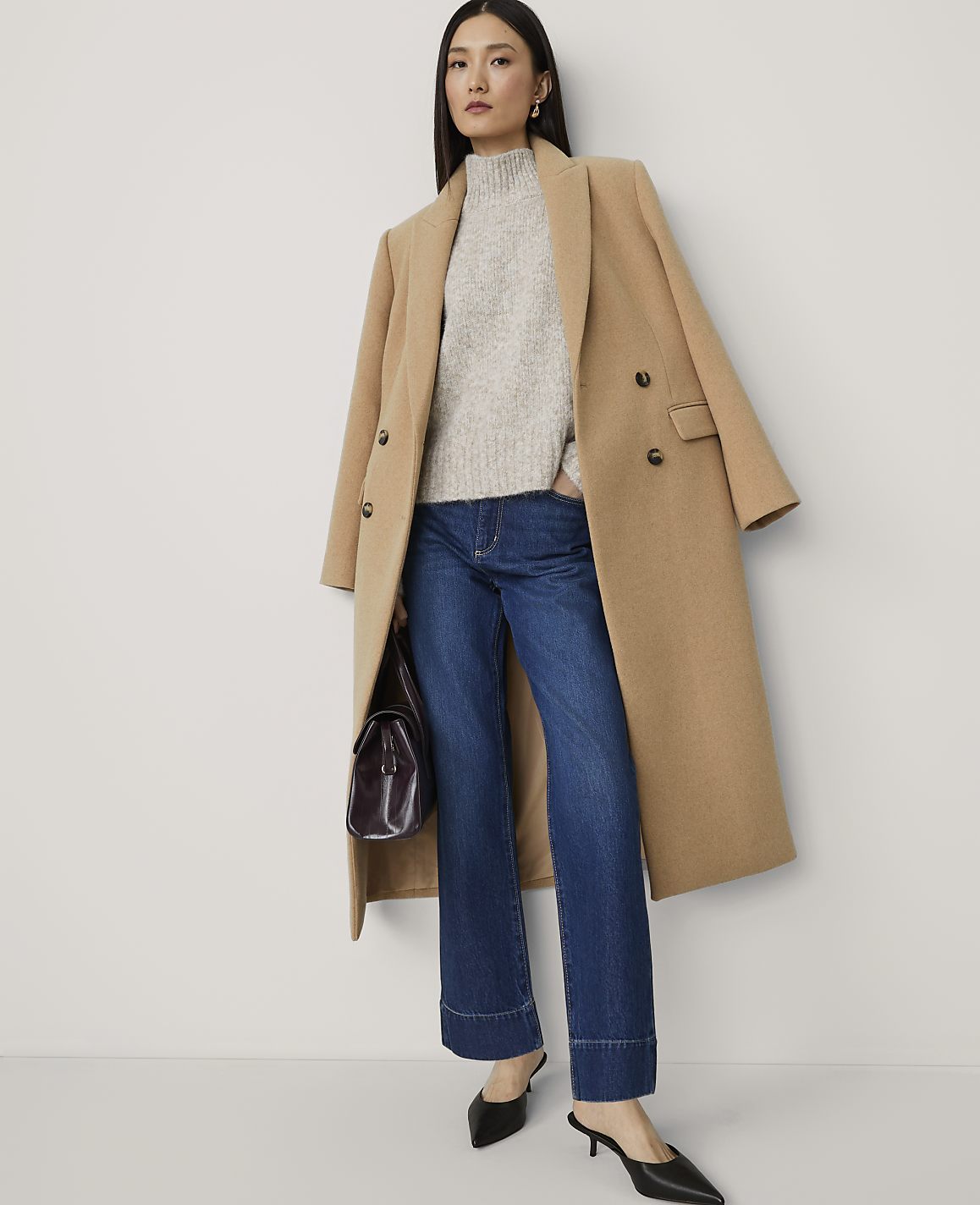 A woman in a camel coat, grey sweater, and wide-leg jeans stands against a plain white background.