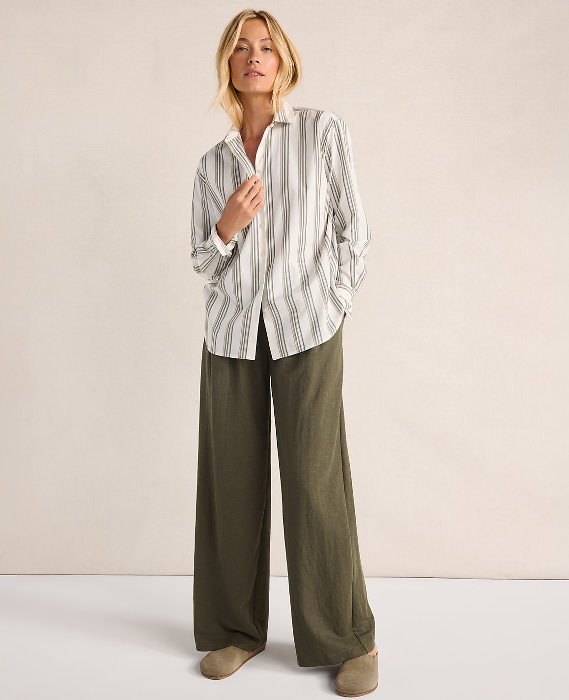 A woman in a striped shirt and wide-leg olive pants stands against a plain, light-colored background.