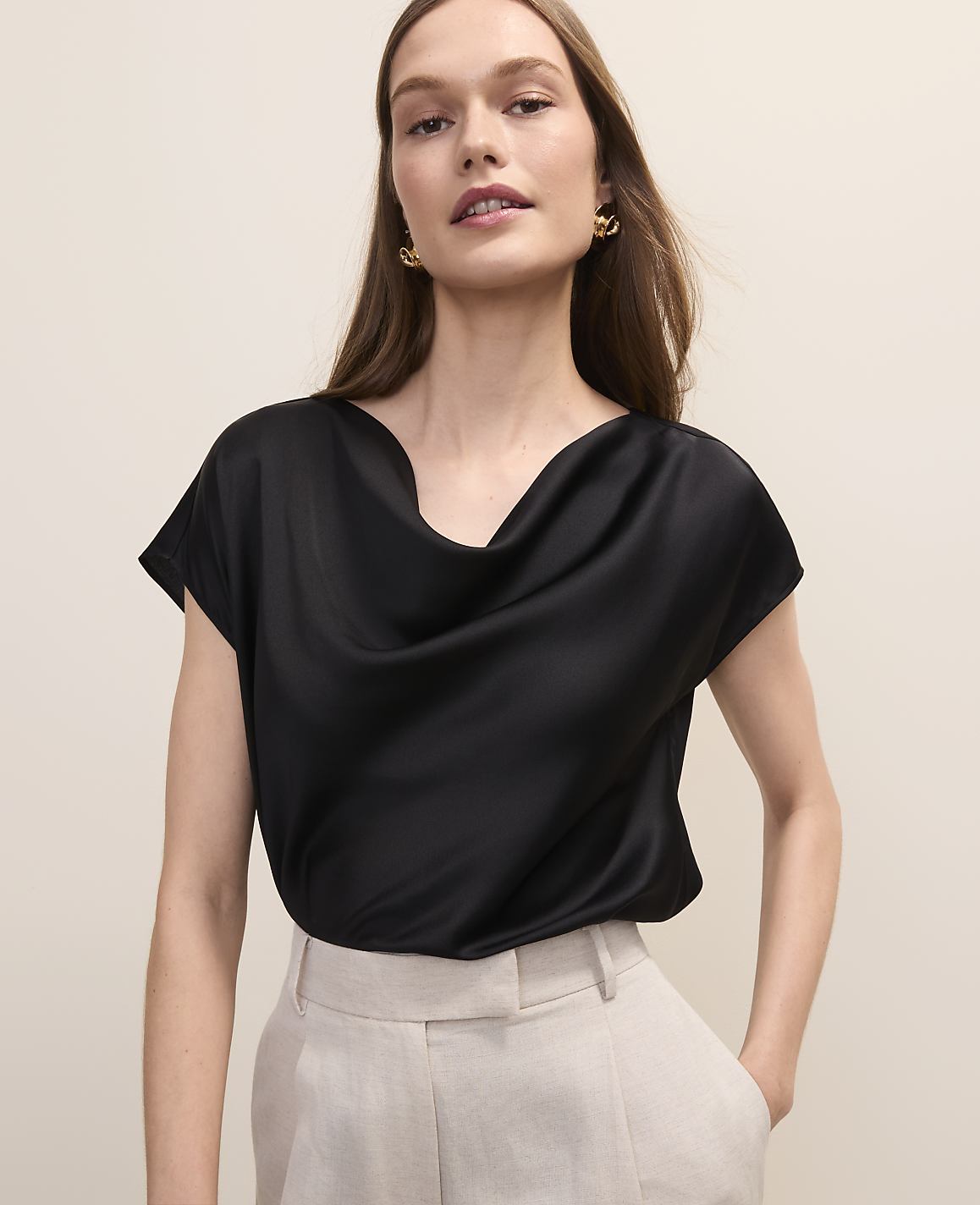 A woman in a black cowl-neck top and beige pants stands against a plain, light-colored background.