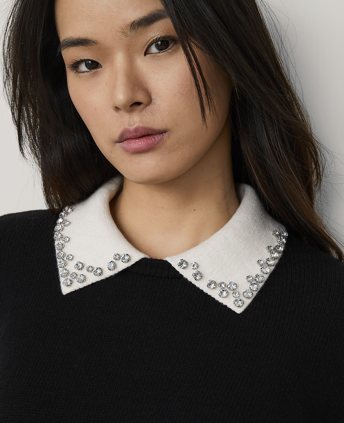 A woman with long dark hair wearing a black sweater with a crystal-embellished white collar poses against a plain light gray background.