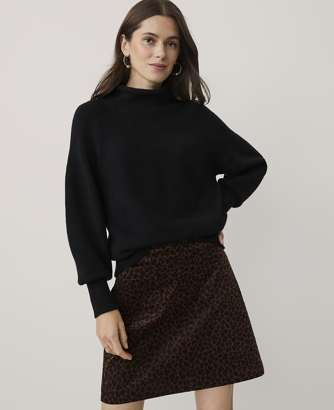A woman wearing a black sweater and a leopard-print skirt stands against a plain, light-grey background.