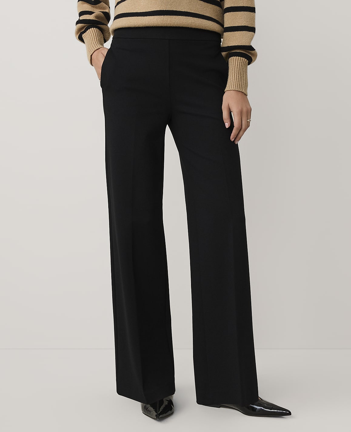 A person wearing a striped tan and black sweater with wide-leg black trousers and pointed black heels stands against a plain light gray background.