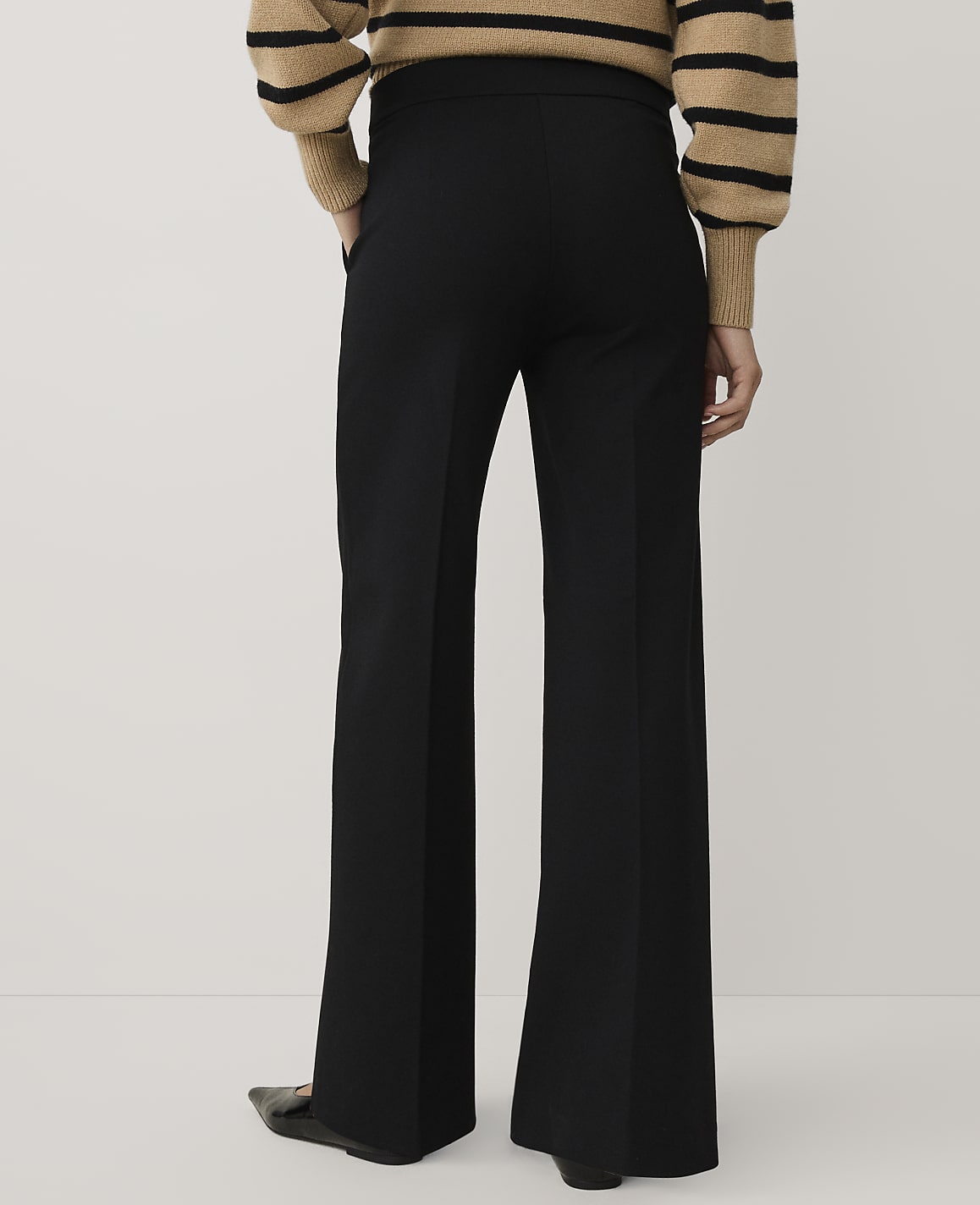 A person wearing a striped tan and black sweater and wide-leg black trousers stands against a plain white background.