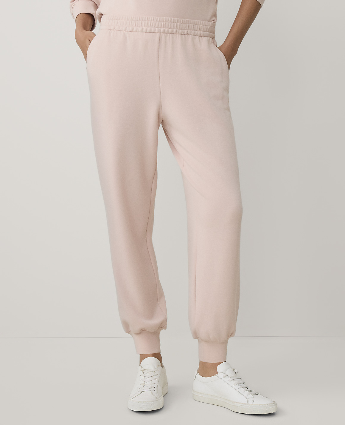 A person wearing light pink sweatpants and white sneakers stands against a plain, light-colored background.