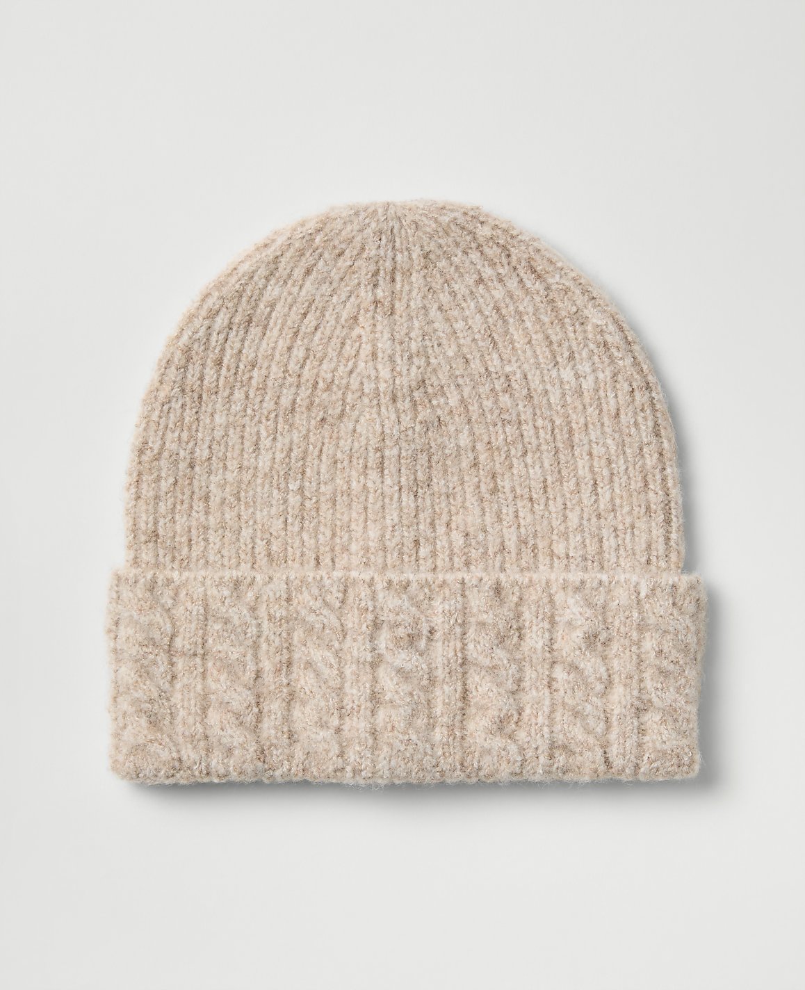 A light beige cable-knit beanie sits centered against a plain, light gray background.
