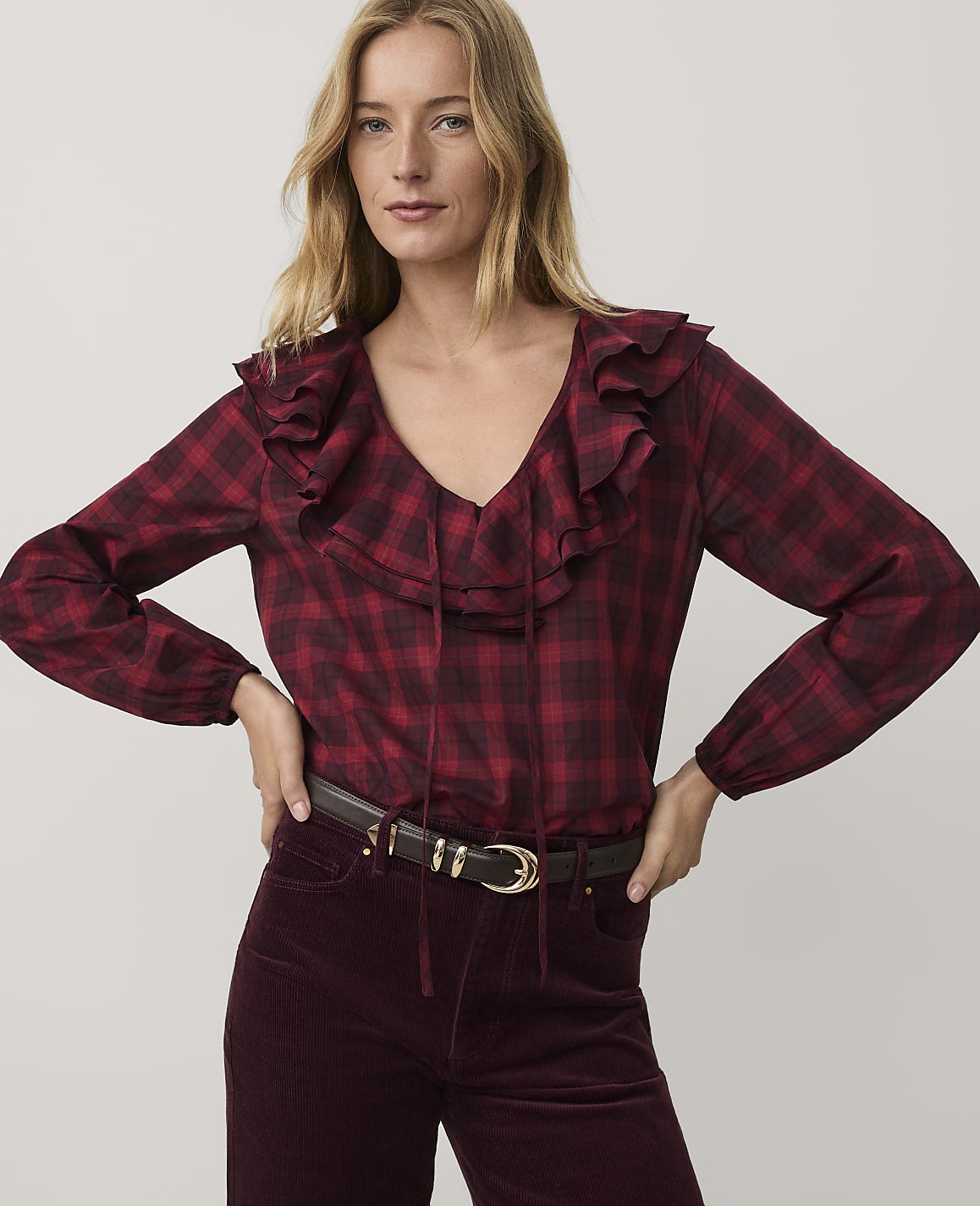 A woman wearing a red plaid ruffled blouse and burgundy corduroy pants stands against a plain light gray background.