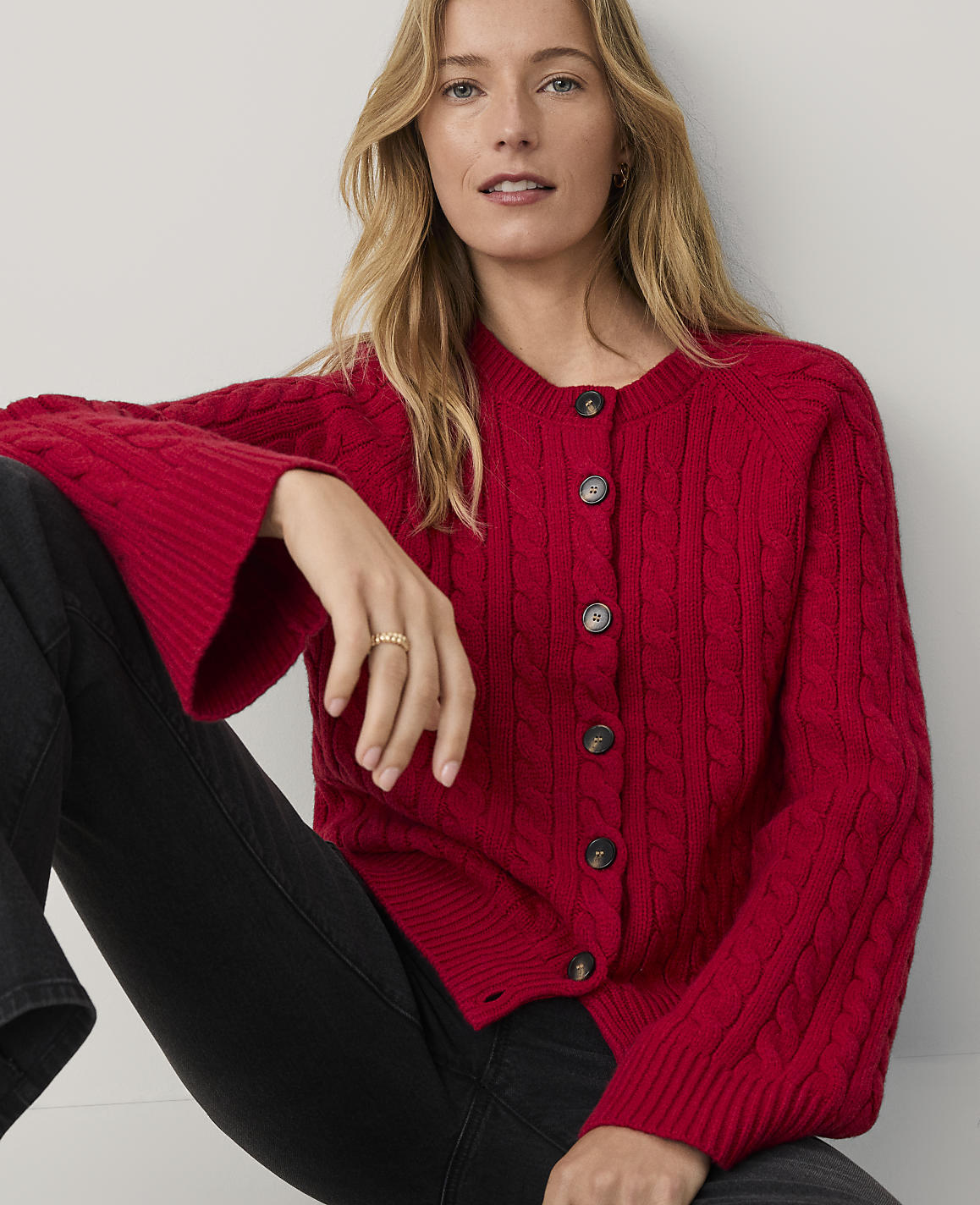 A blonde woman wearing a red cable-knit cardigan and black pants sits against a plain white background.