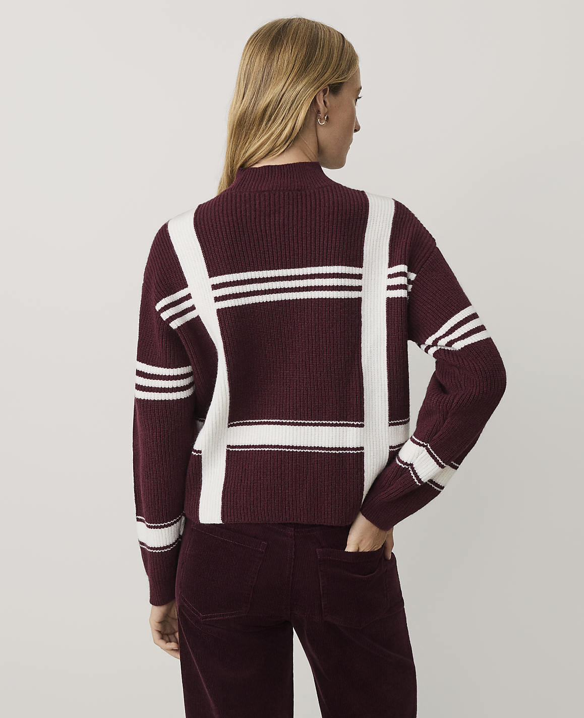 A woman wearing a burgundy and white plaid sweater and matching corduroy pants stands against a plain light gray background.