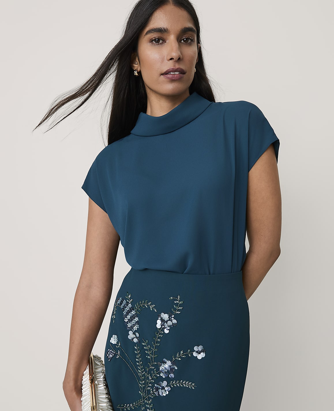 A woman wearing a teal two-piece outfit with floral sequins stands against a plain, light-colored background.
