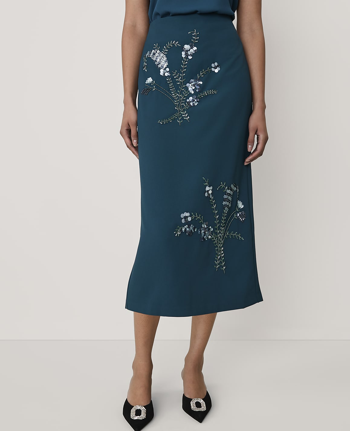 A person wears a teal midi skirt with floral sequin embroidery and black pointed-toe heels against a plain white background.