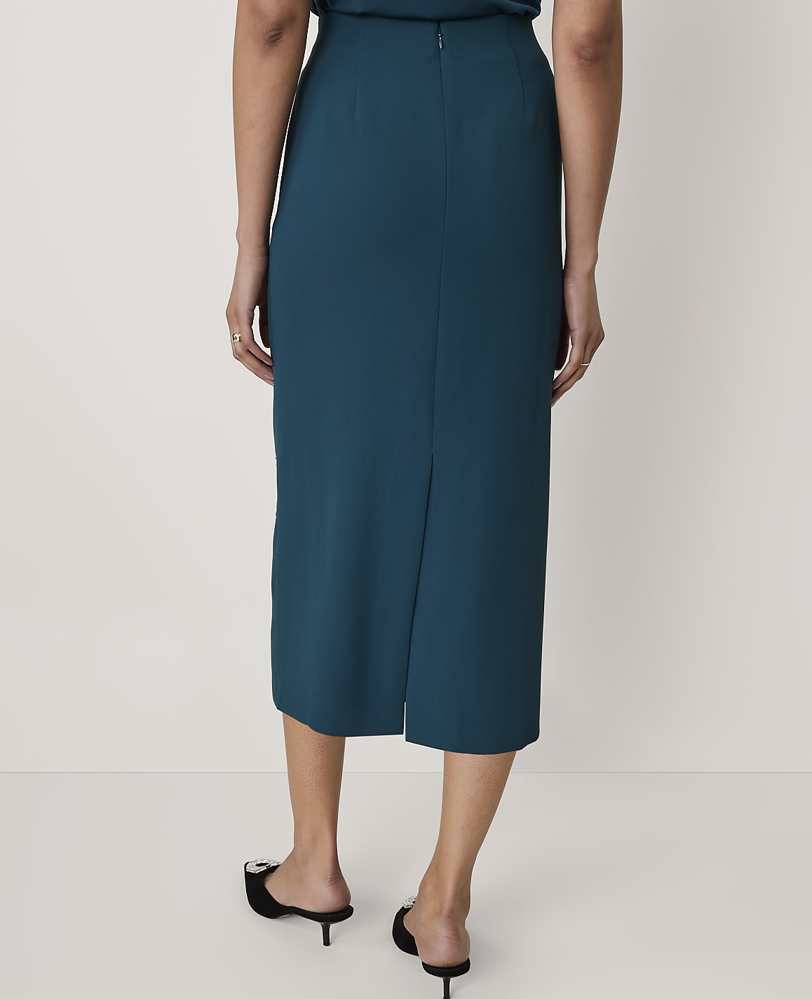 A person wearing a teal midi skirt and black pointed-toe mules stands against a plain white background.