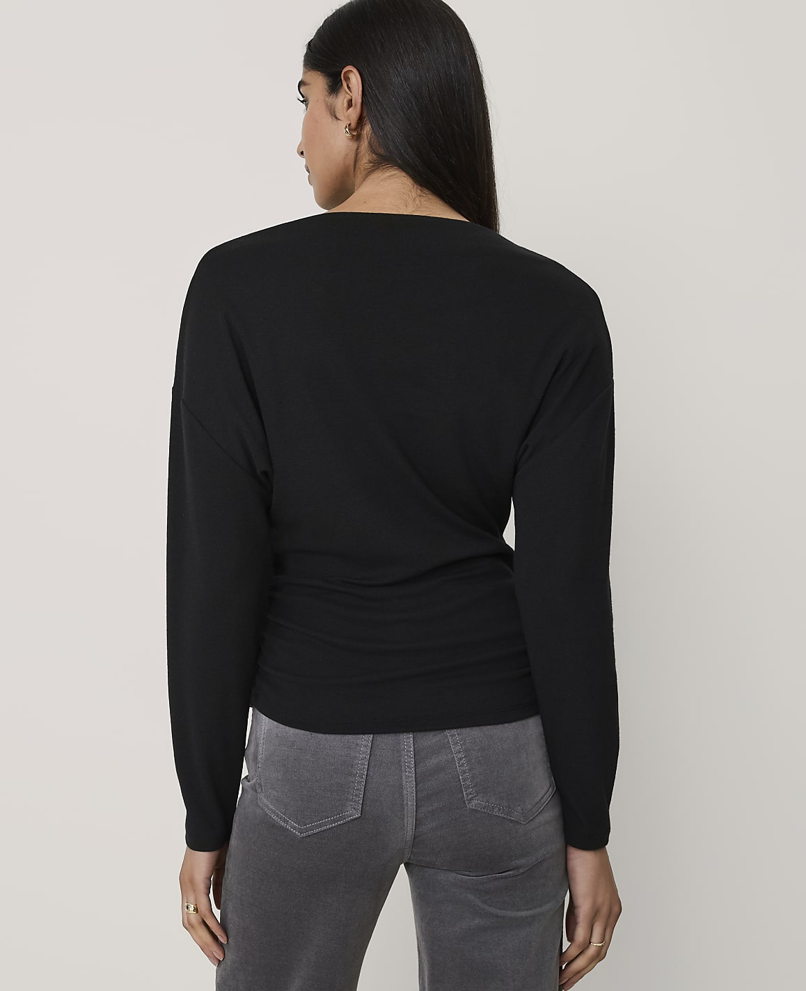 A woman wearing a black long-sleeved top and grey pants stands with her back to the camera against a plain light grey background.