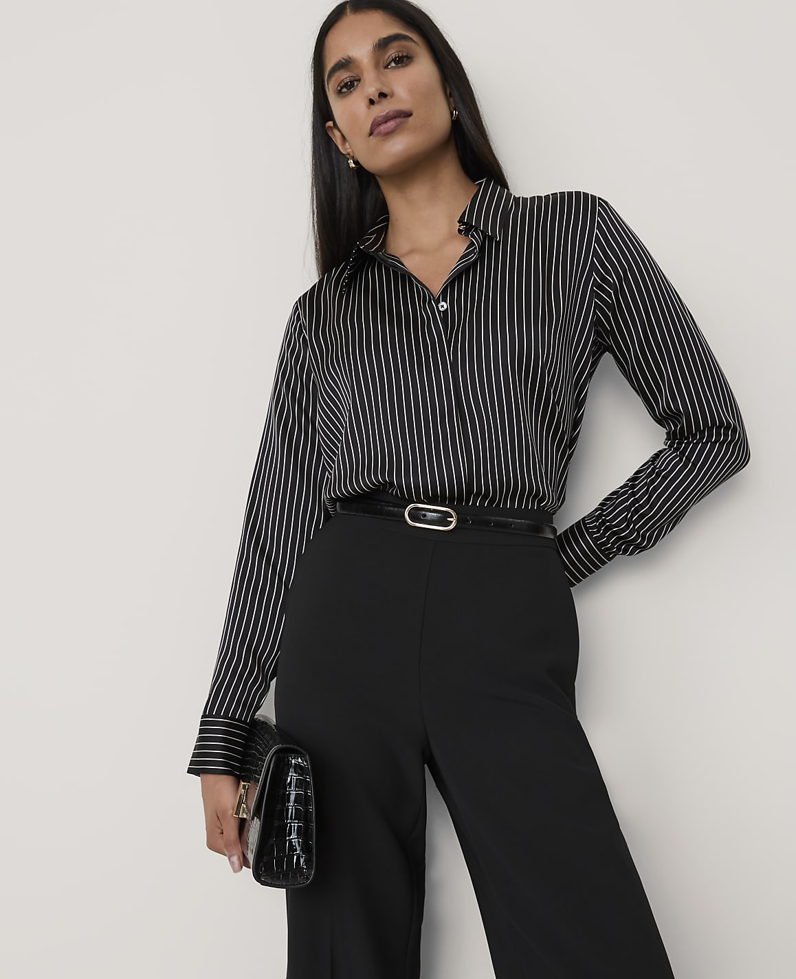 A woman wearing a black and white striped shirt and black trousers stands against a plain white background.