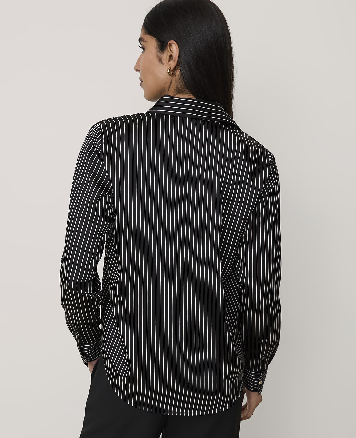 A woman wearing a black and white pinstriped shirt and black trousers stands against a plain light gray background.