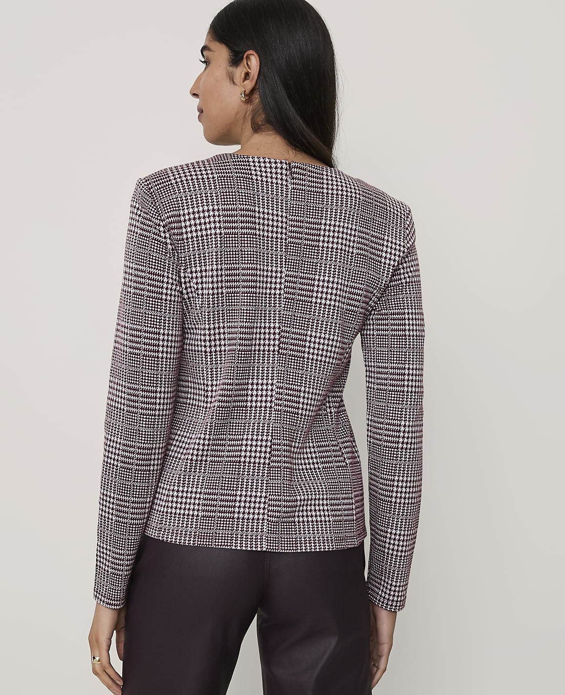 A woman stands with her back to the camera wearing a maroon and white houndstooth blazer and dark trousers against a plain light grey background.