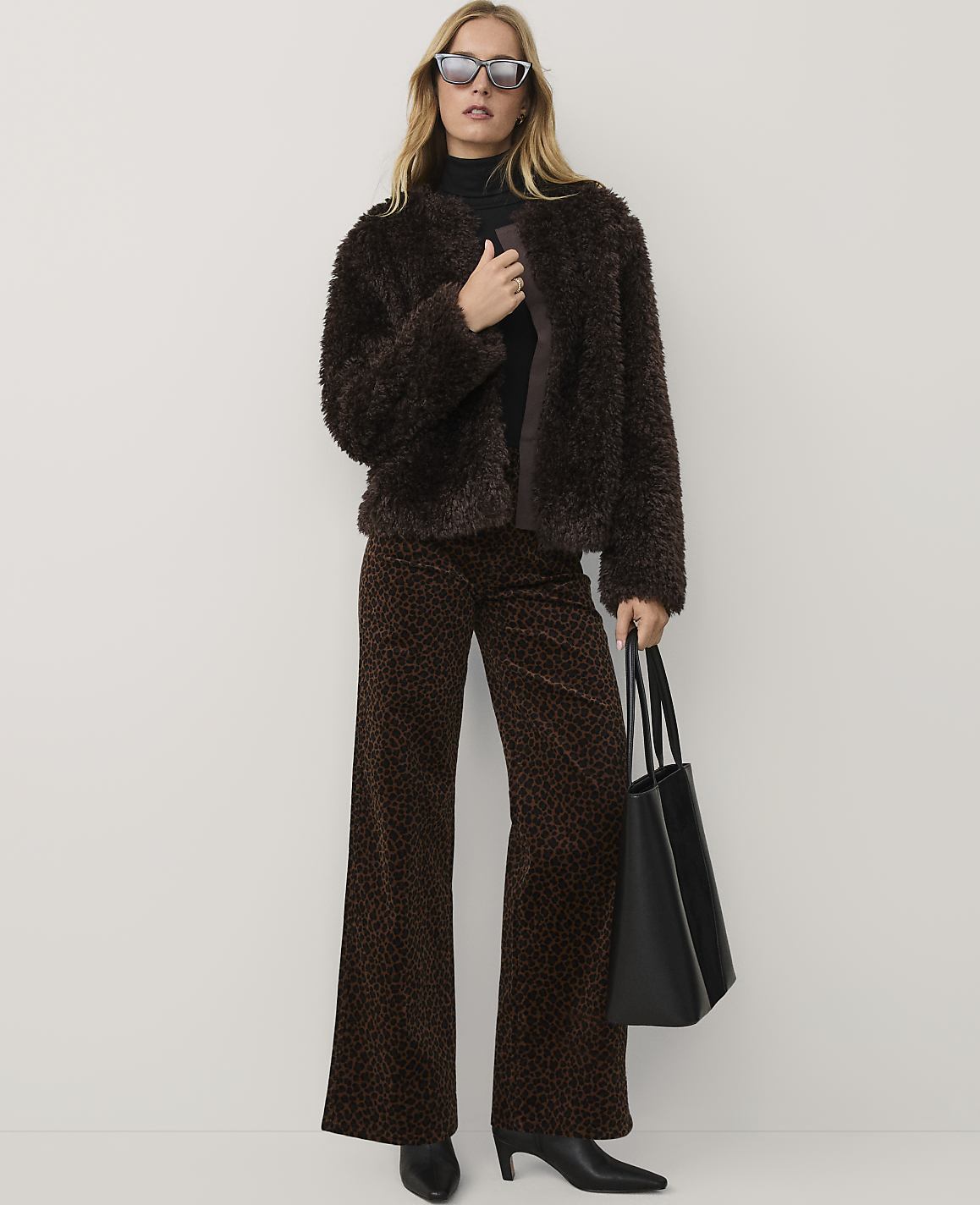 A woman wearing a brown faux-fur jacket, leopard-print trousers, and sunglasses stands against a plain white background.