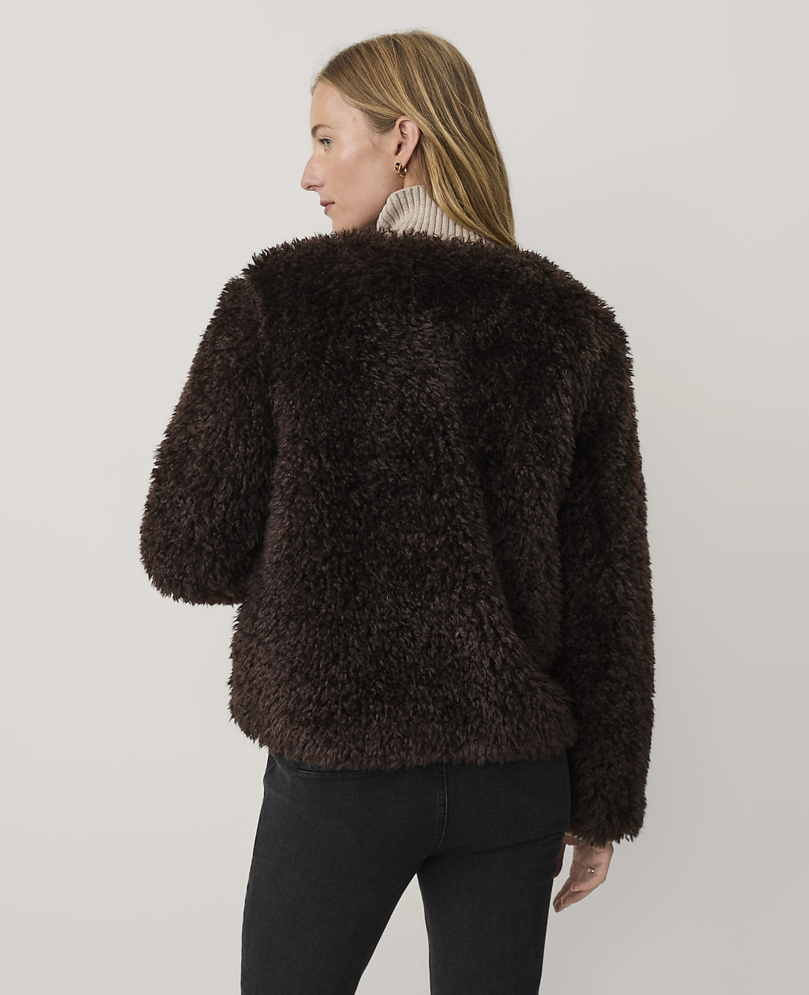 A woman wearing a dark brown faux-fur jacket and black jeans stands against a plain, light-colored background.