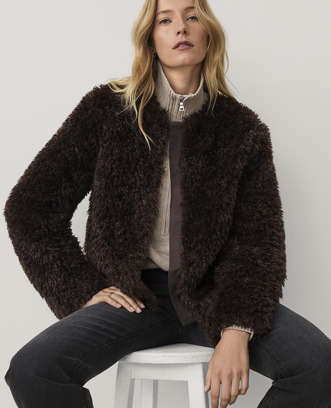 A woman wearing a dark brown faux-fur jacket and black jeans sits on a white stool against a plain light-colored background.