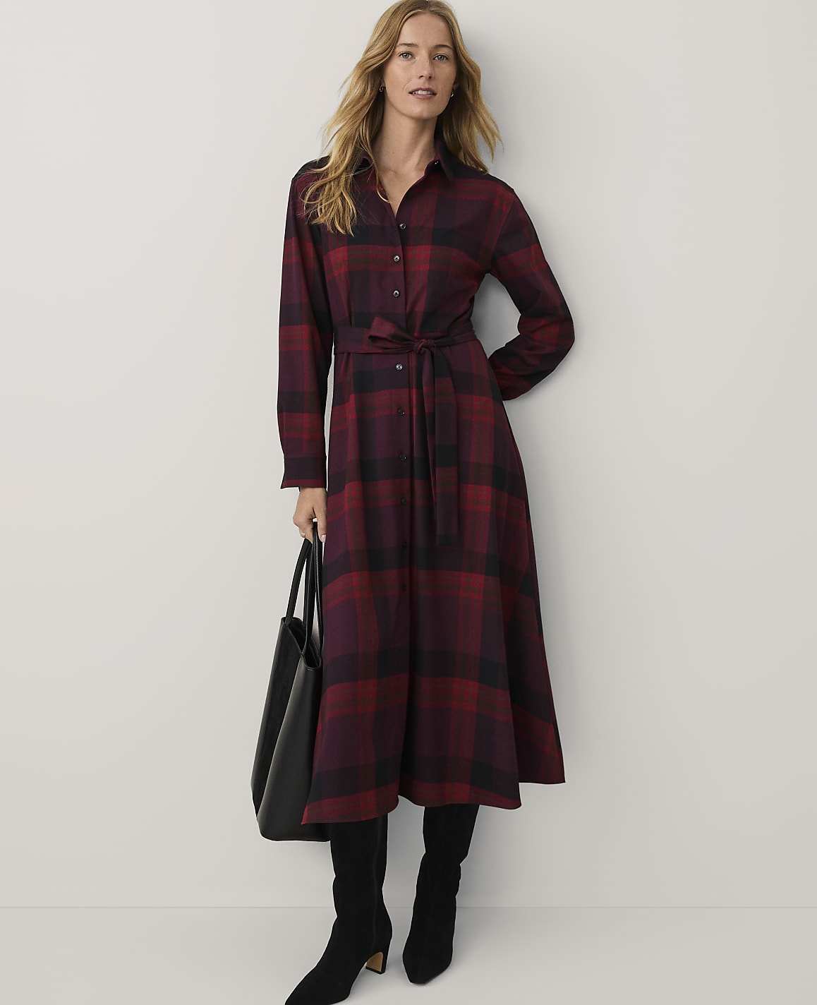 A woman wearing a red and black plaid maxi dress and black boots stands against a plain white background while holding a black handbag.