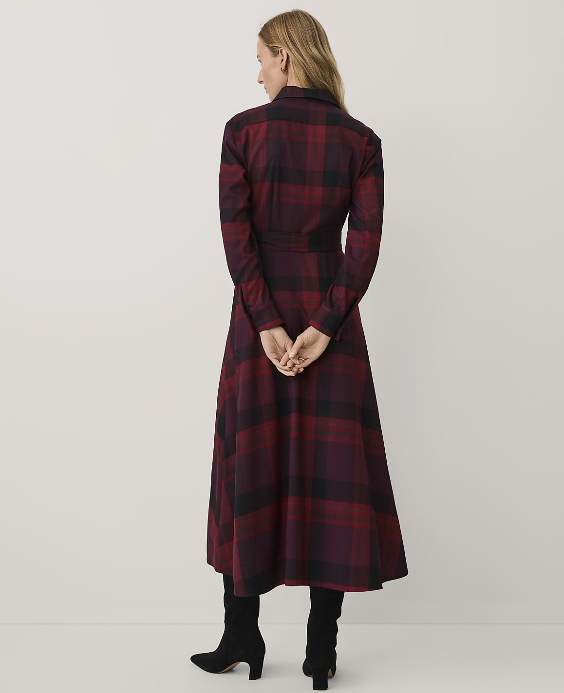 A woman wearing a red and black plaid dress and black boots stands with her back to the camera against a plain white background.