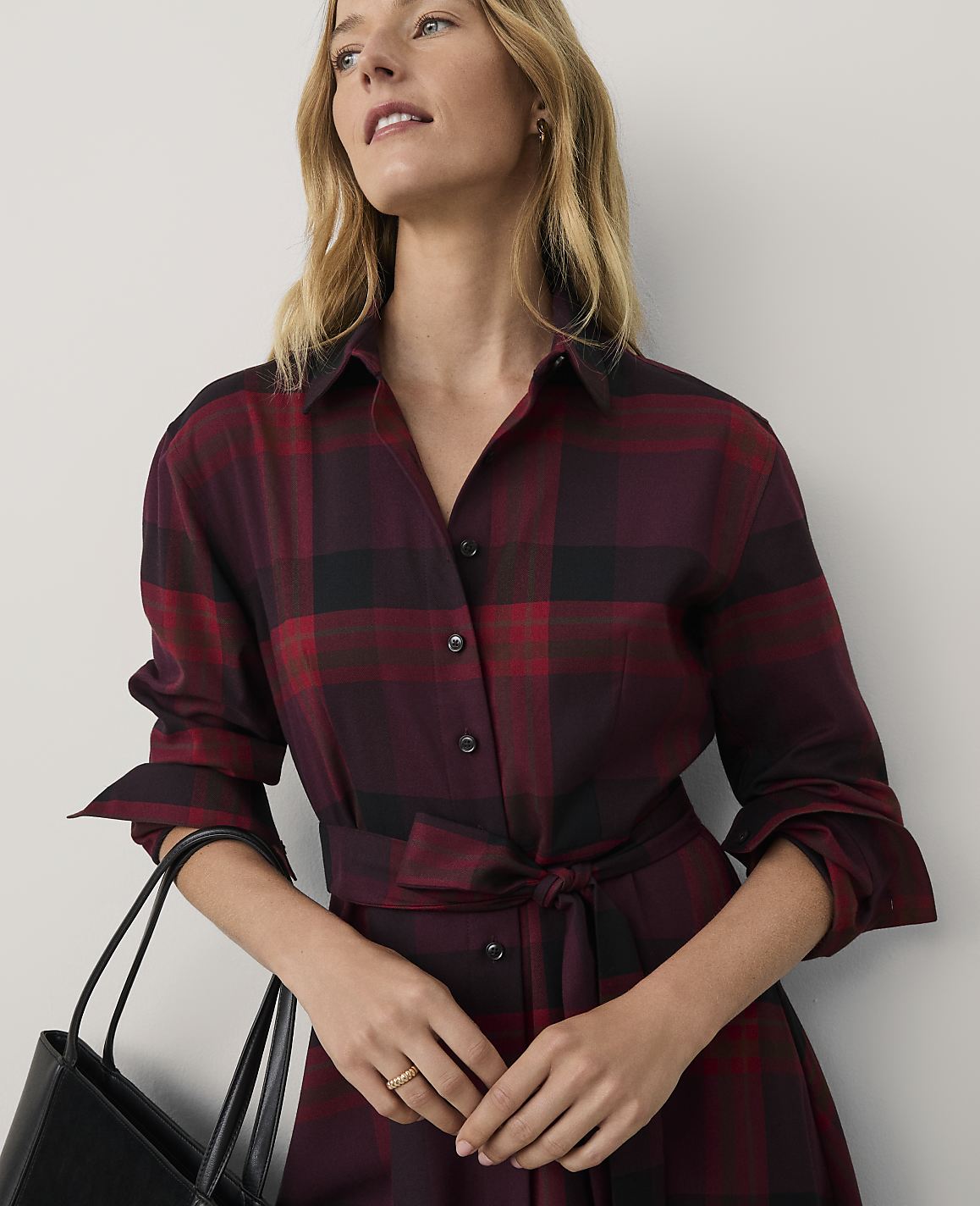 A woman wearing a red and black plaid dress holds a black handbag against a plain white background.