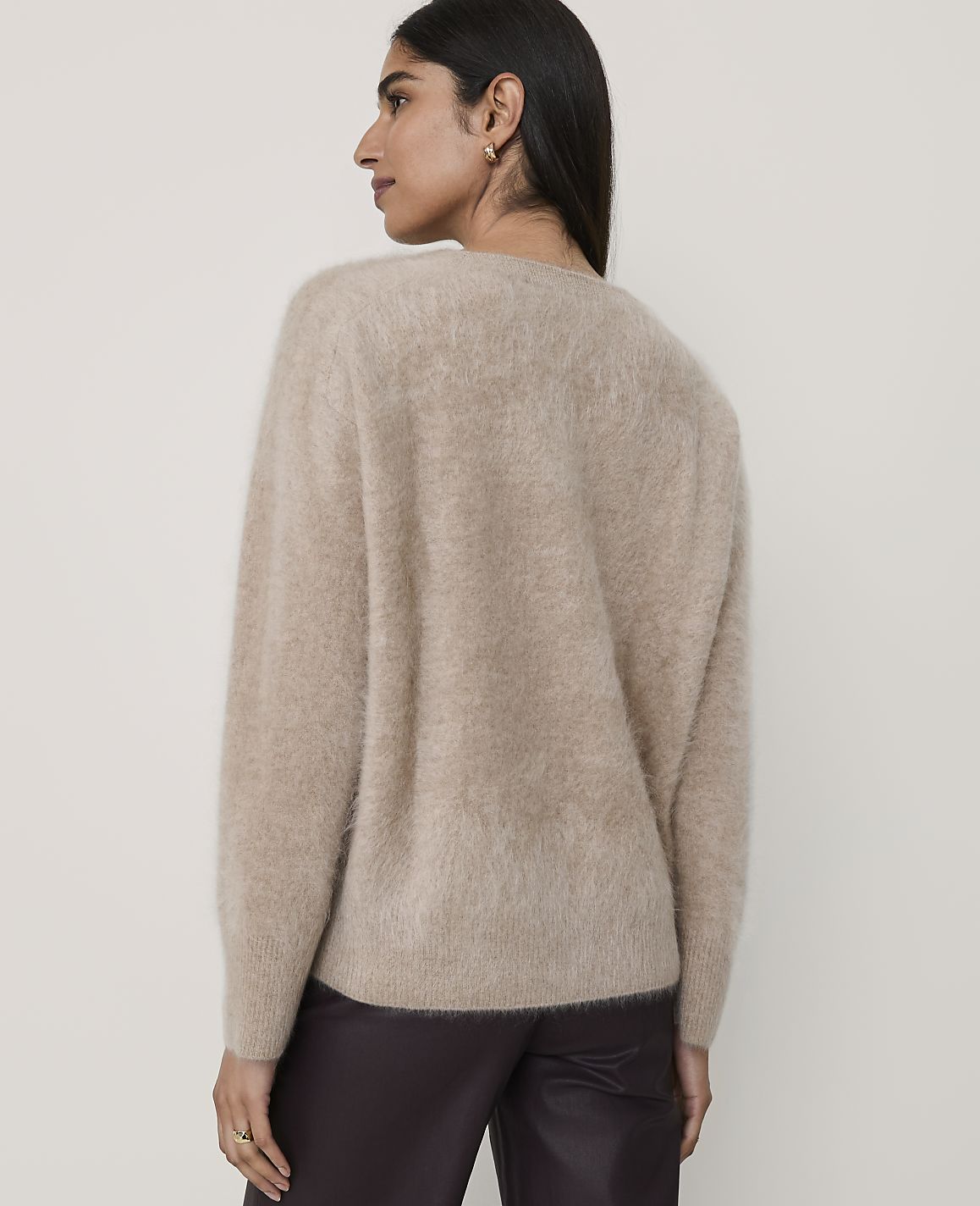 A woman wearing a fuzzy beige sweater and dark trousers stands against a plain, light-colored background.