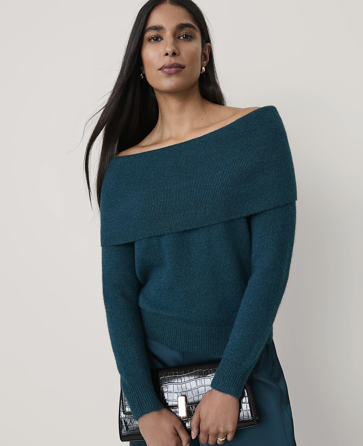 A woman wearing a teal off-the-shoulder sweater and holding a black crocodile-patterned clutch poses against a plain, light-colored background.