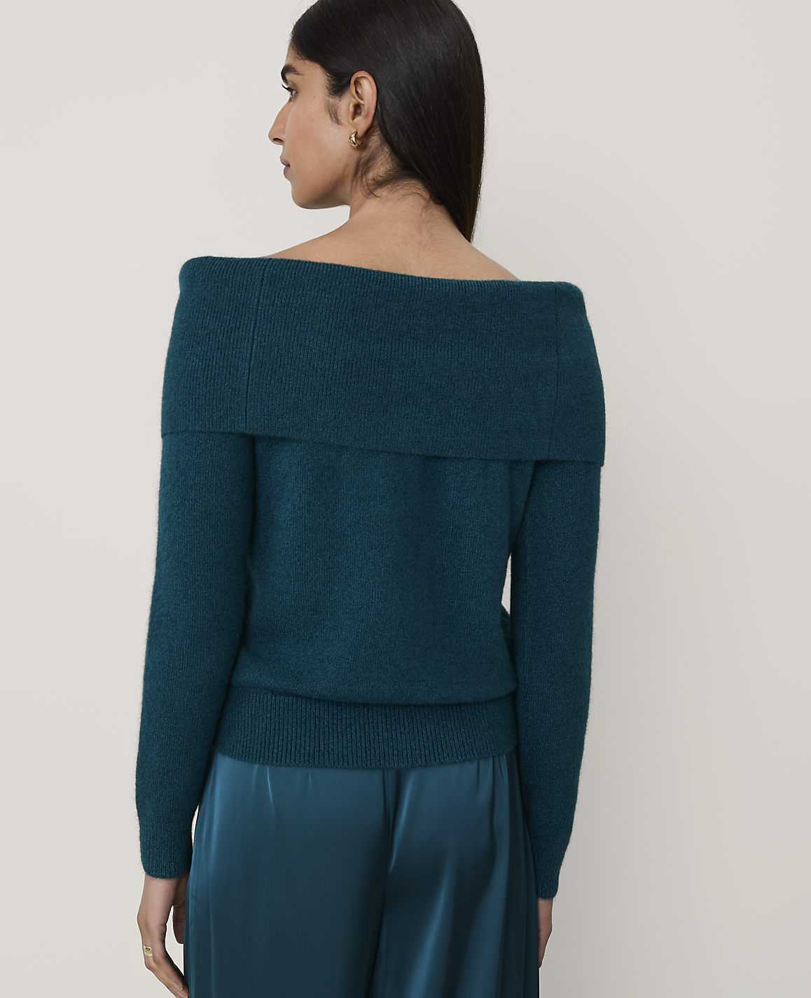 A woman wearing a teal off-the-shoulder sweater and matching satin skirt stands against a plain, light-colored background.