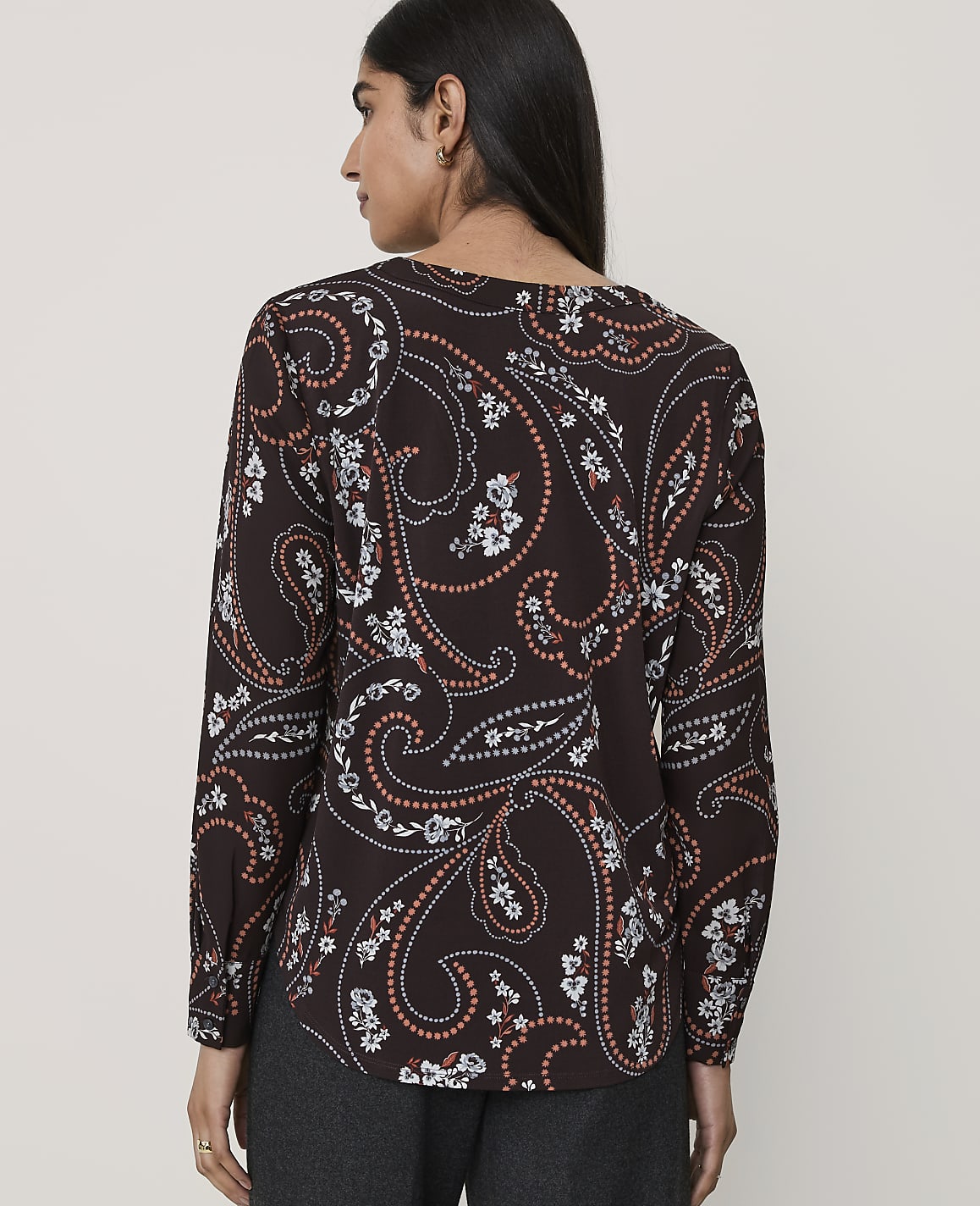 A woman wearing a brown paisley-print blouse and dark trousers stands against a plain, light-colored background.